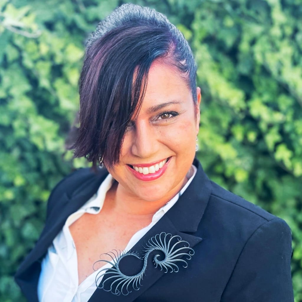 A person with shoulder-length dark hair, wearing a black blazer over a white shirt, smiles at the camera. The lush green foliage in the background hints at the celebratory atmosphere of the MassArt Common Good Awards.