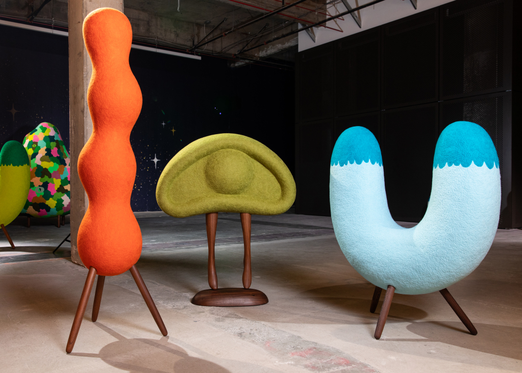 Three colorful, abstract sculptural chairs with wooden legs sit in a modern, industrial indoor space at the MassArt Art Museum’s 2026 Contemporary Art Exhibitions. The orange, green, and blue chairs feature unique, whimsical shapes.
