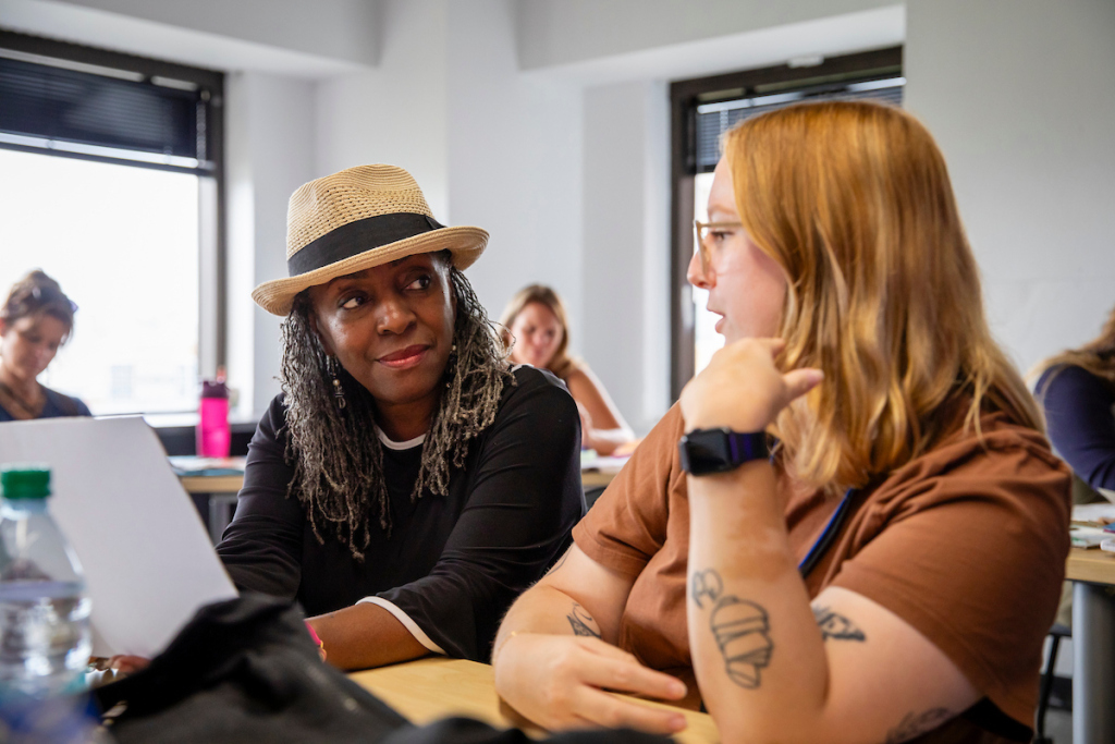 Two women sit at a table in a classroom, engaged in conversation during Admissions Meetings. One wears a hat and black top, while the other has blonde hair, glasses, and tattoos. Other people work in the background.