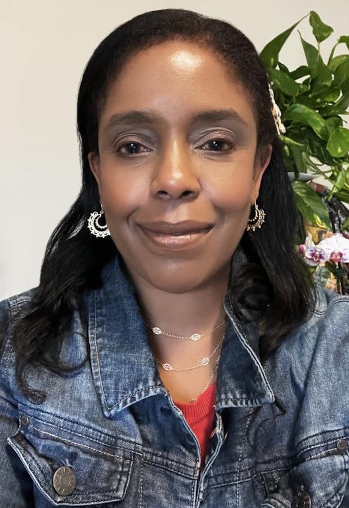A woman with long dark hair smiles softly at the camera, wearing a denim jacket, layered necklaces, and hoop earrings. There is a green plant in the background.