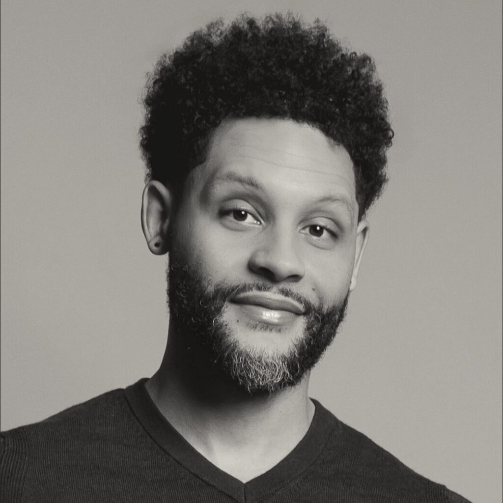A man with curly hair and a beard smiles softly at the camera. Wearing a dark V-neck shirt and a small earring, he appears in black and white against a plain background, evoking the creative spirit of MassArt's Common Good Awards.