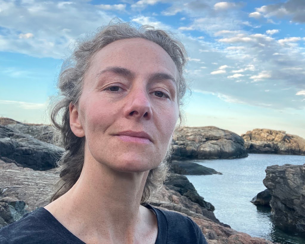 A person with wavy gray hair stands on rocky terrain near the sea, with calm water, large rocks, and a partly cloudy blue sky in the background.