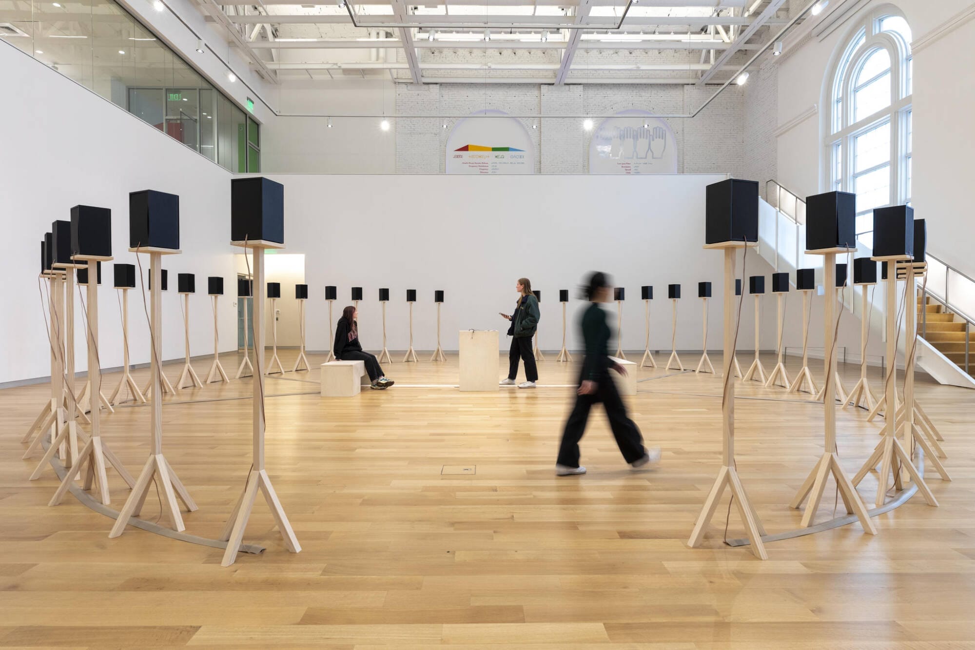 Visitors explore a student and faculty art exhibition in a bright MassArt gallery with large windows and high ceilings.