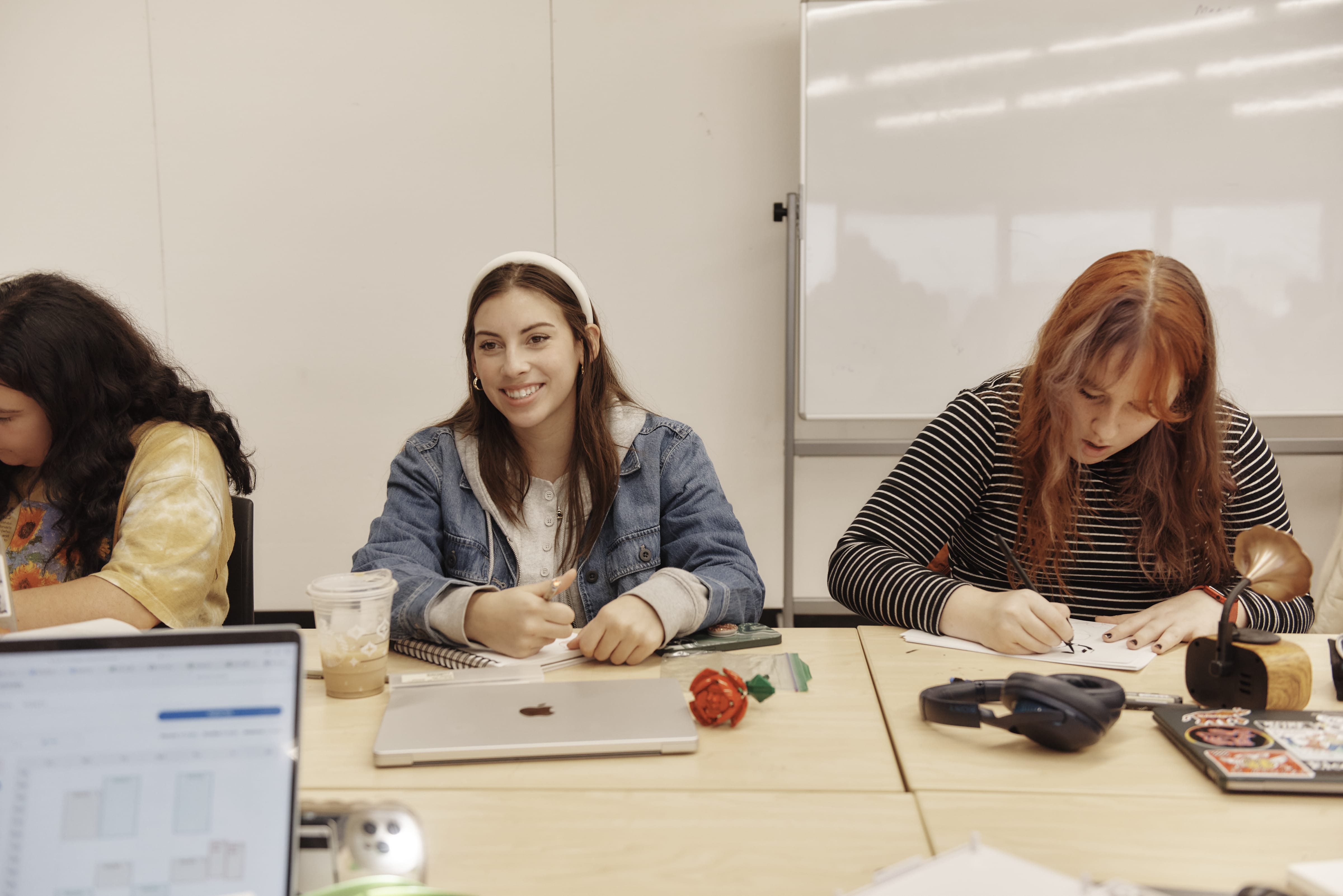 Three students sit at a table in a classroom. One smiles and holds a pencil, another writes in a notebook, and the third looks down—perhaps reviewing MassArt admissions requirements or their MassArt application. A laptop and coffee are on the table; a whiteboard is behind them.