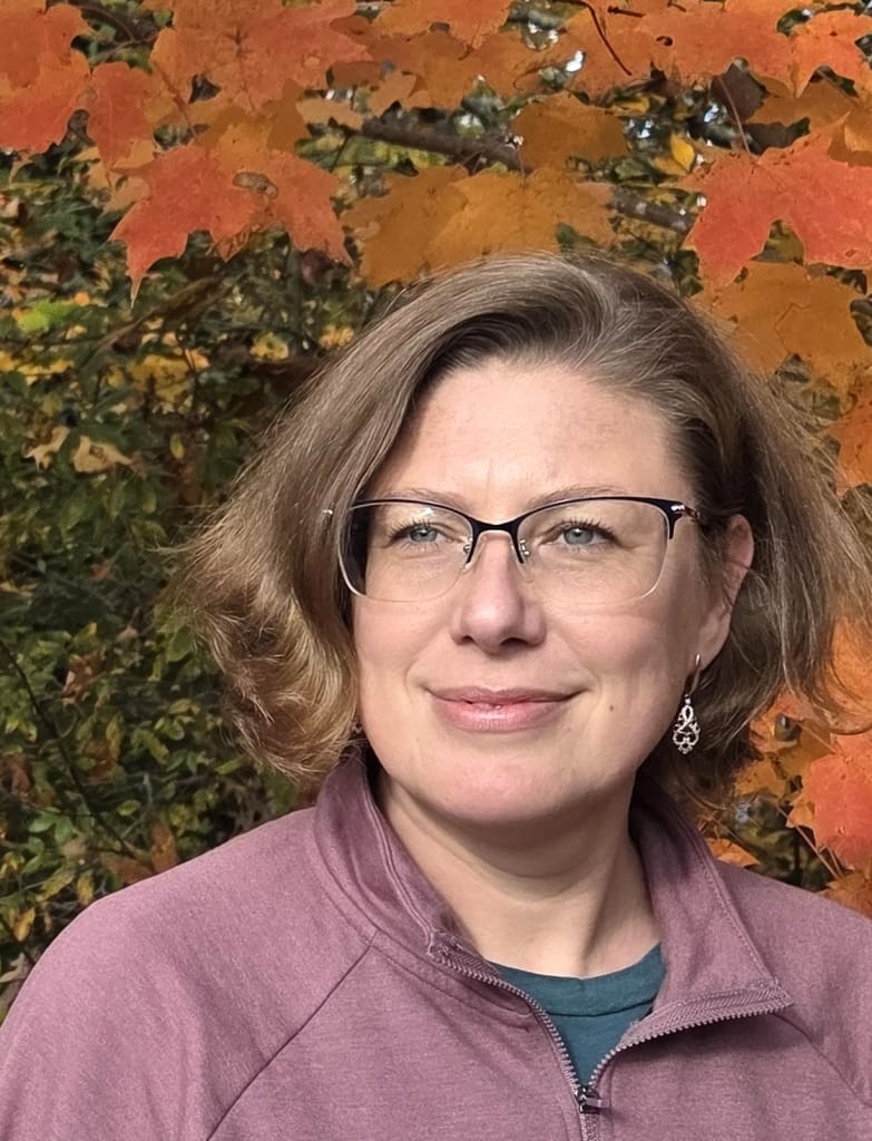 A woman with short light brown hair and glasses stands outdoors, wearing a maroon zip-up jacket. Behind her are vibrant orange autumn leaves and green foliage. She is smiling softly and looking slightly to the right.