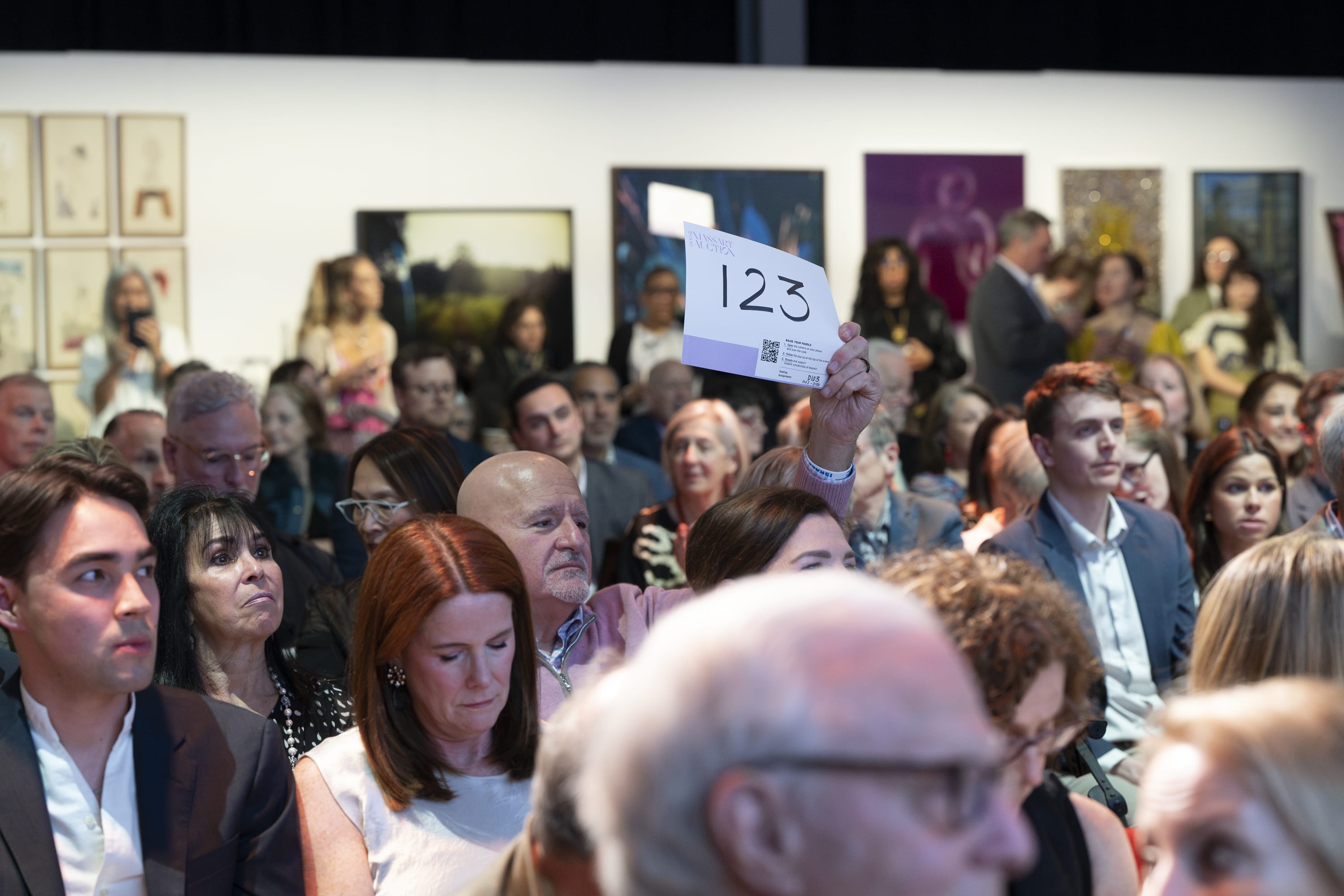 A person seated in a crowded auction holds up a card with the number 123. People around, including sponsors, are seated and watching attentively. Framed artwork is visible on the walls in the background.