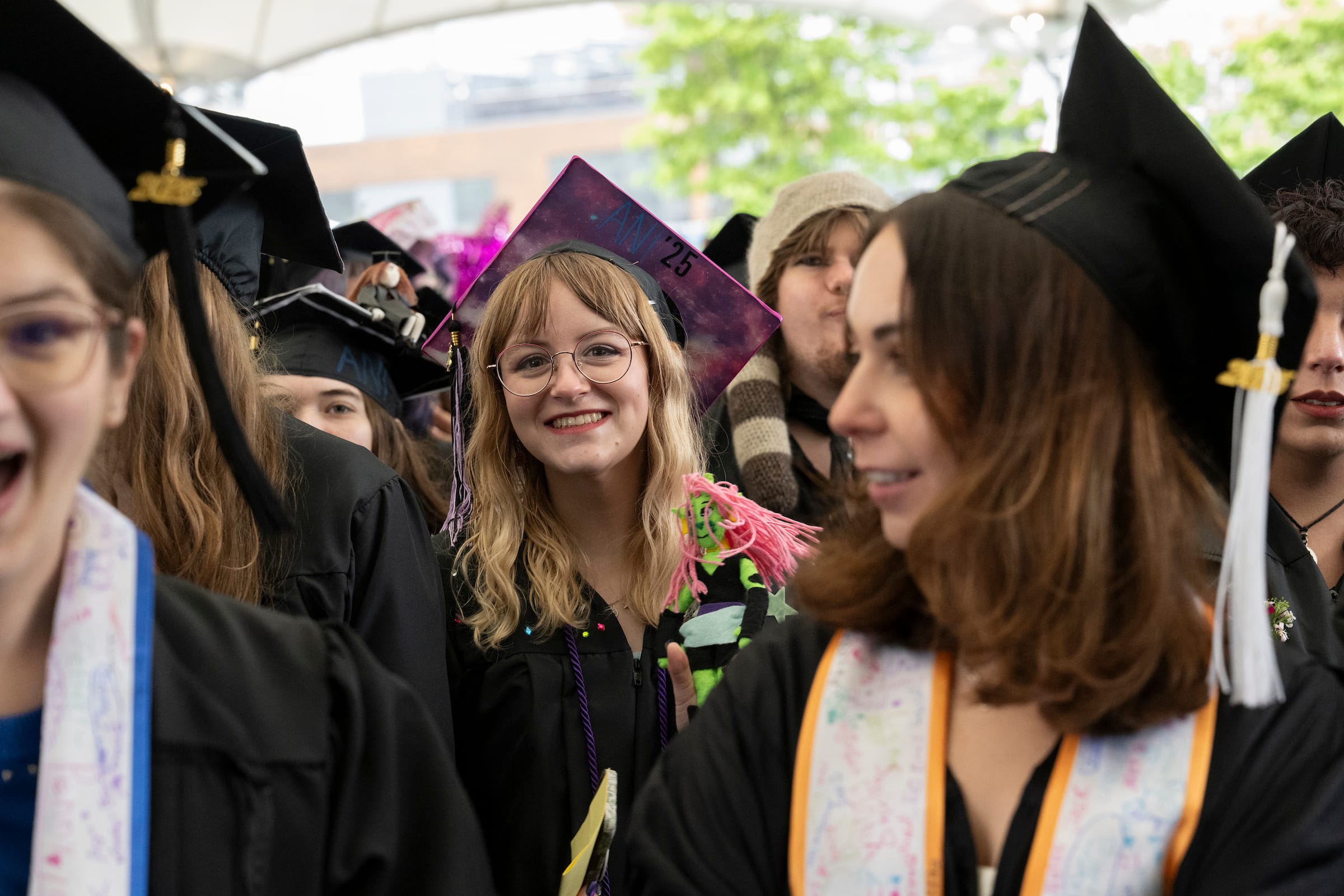 A group of graduates in caps and gowns smile and celebrate at an outdoor commencement ceremony. One person in the center, wearing glasses, smiles directly at the camera while holding decorated graduation caps.
