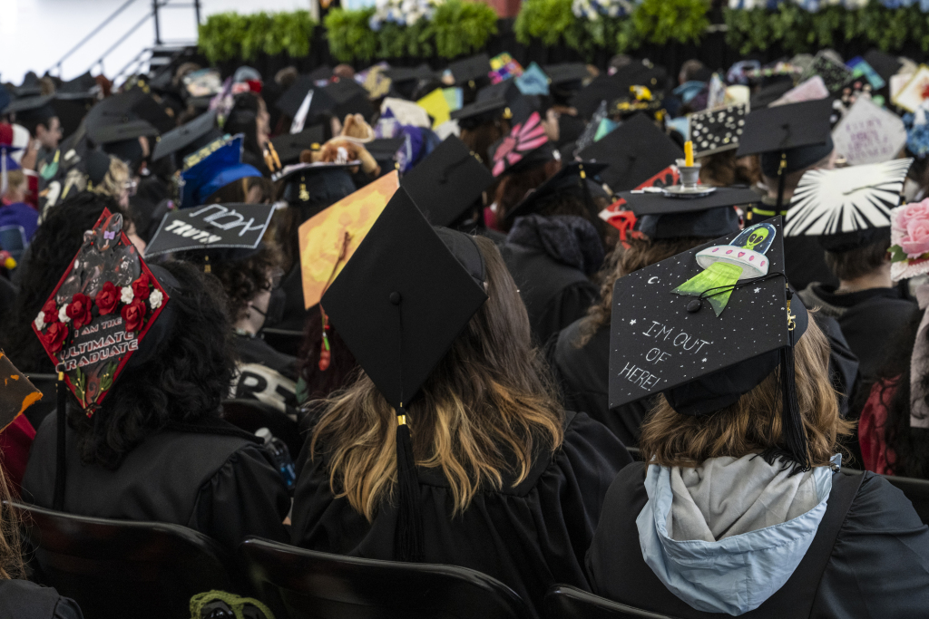 A group of graduates sit in rows at a lively commencement, wearing decorated caps with colorful designs like a UFO proclaiming "I'm out of here!" and flowers. The setting appears to be an indoor graduation ceremony.