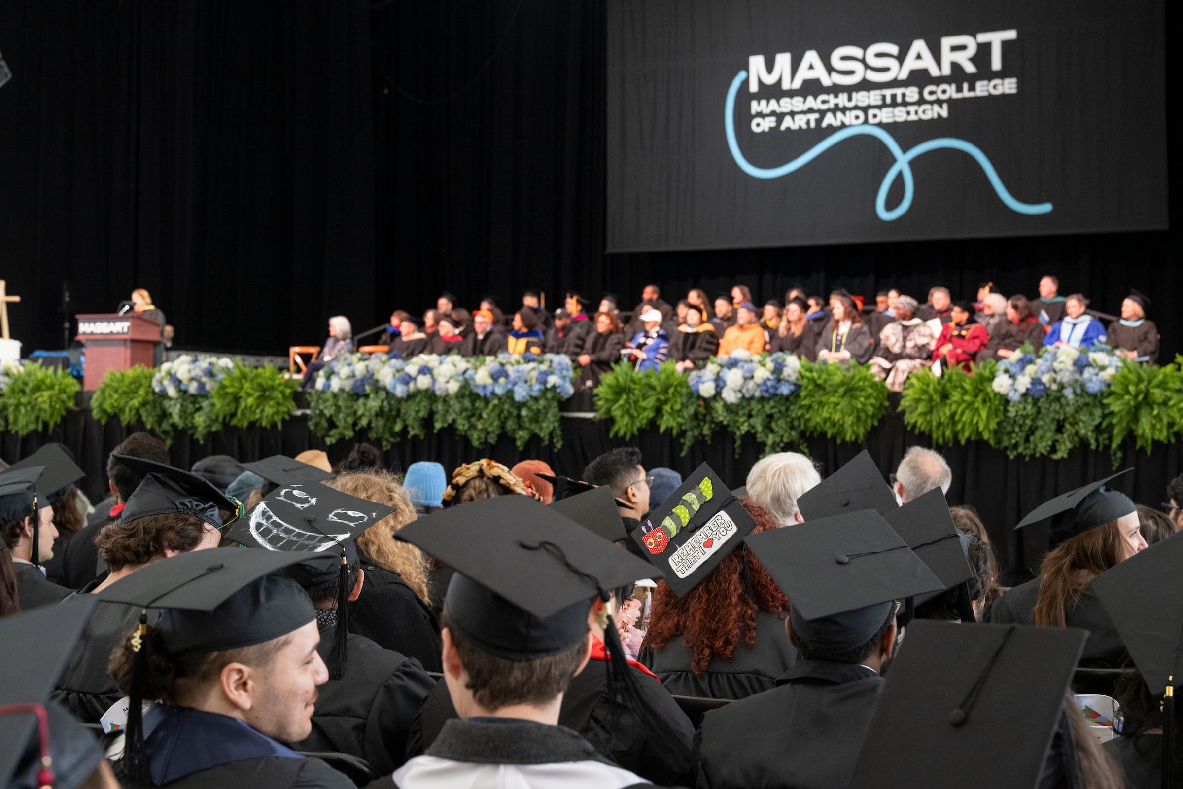 A commencement ceremony at MassArt features graduates in caps and gowns seated in the audience, while faculty and speakers sit on stage behind a podium, with the MassArt logo displayed on a screen above them.
