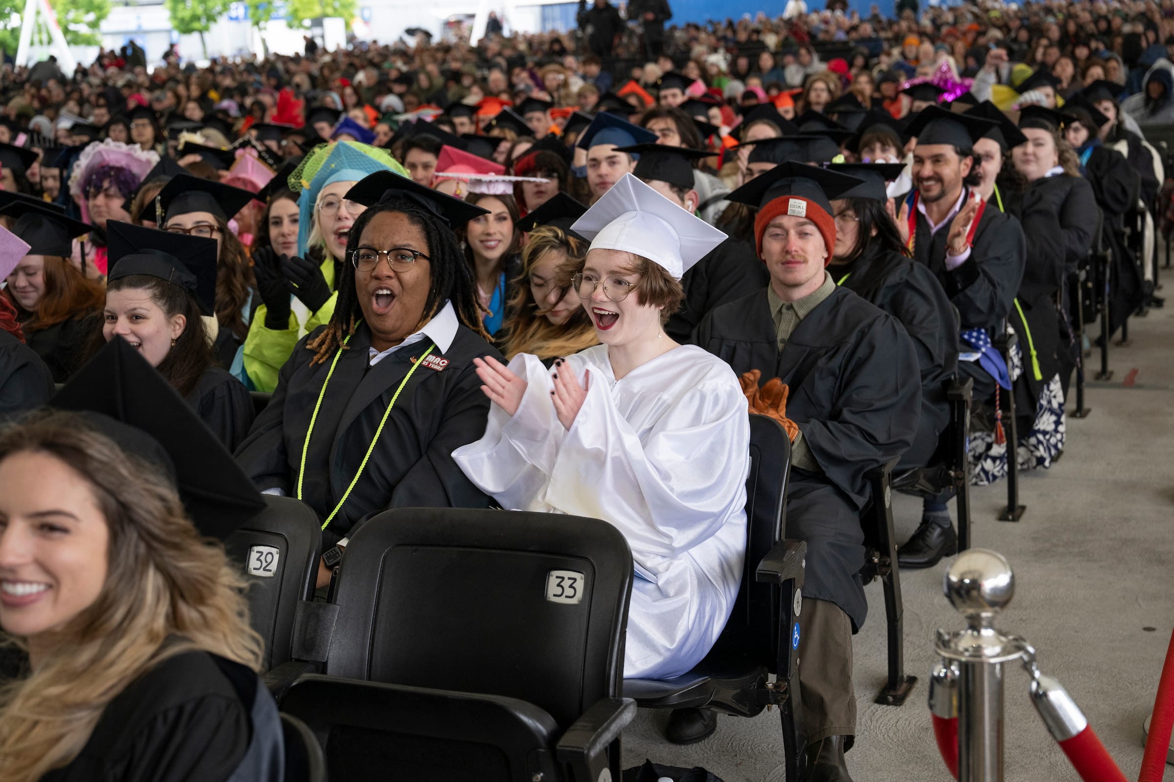 Rows of graduates in caps and gowns sit together at a commencement ceremony. One graduate in a white gown sits among others in black gowns, clapping and smiling, while the crowd around them cheers in celebration of graduation.