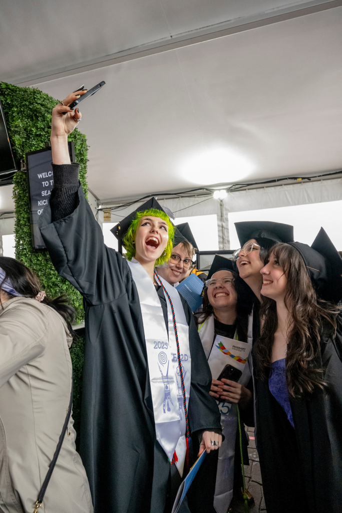 A group of graduates in caps and gowns smile and pose for a selfie at a commencement ceremony under a tent, with one person holding up a phone and others gathering closely.