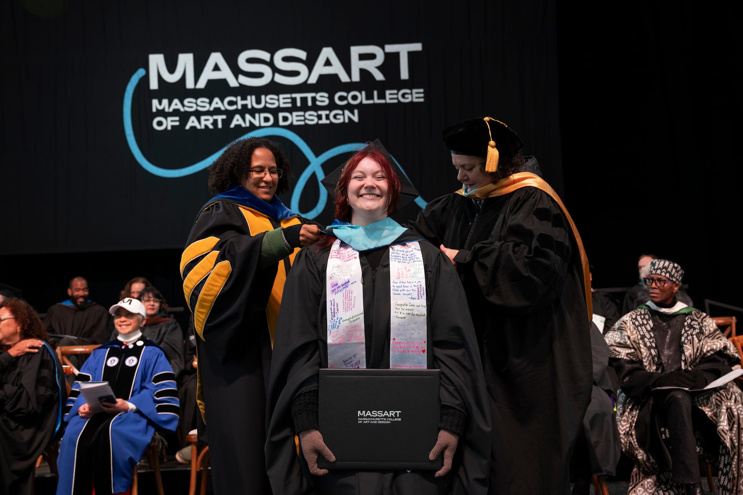 A smiling graduate in cap and gown is hooded by two faculty members on stage at Massachusetts College of Art and Design, with the college’s logo projected in the background during a joyful commencement ceremony.