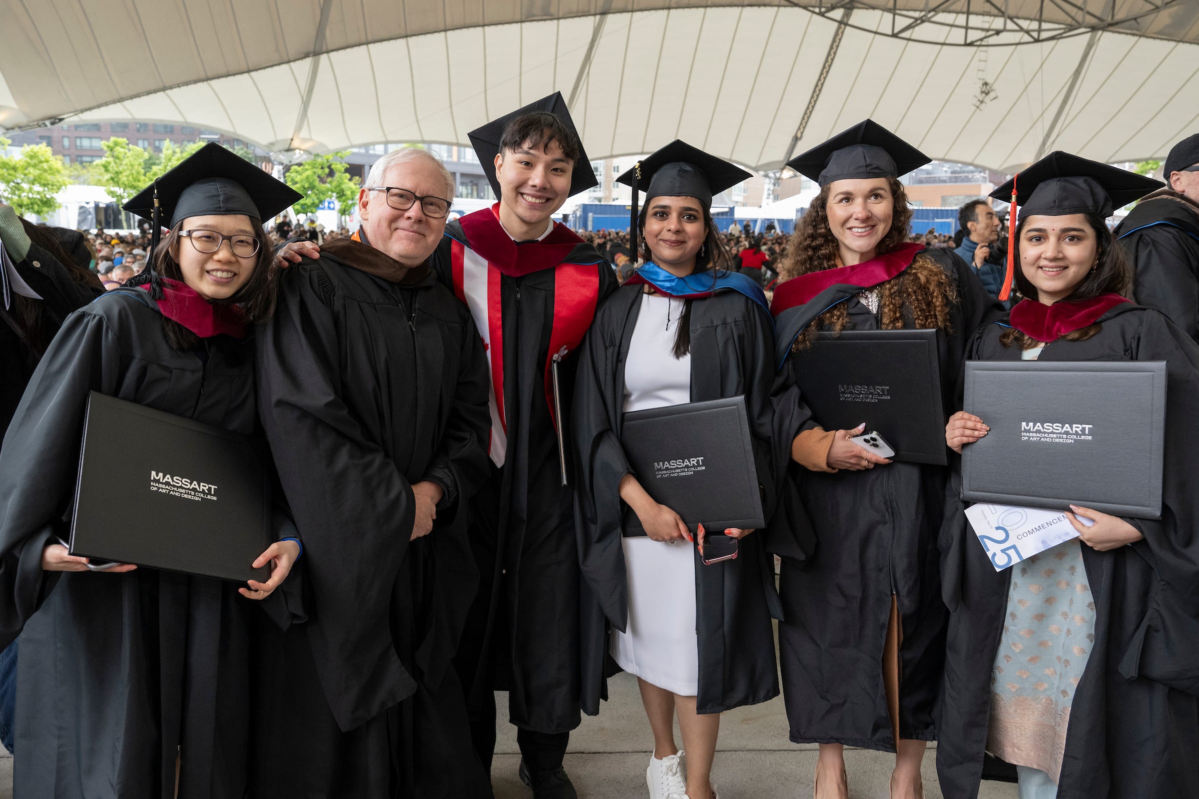 Six graduates in caps and gowns smile and pose together at a commencement ceremony, proudly holding their diplomas under a large tent with people gathered in the background.