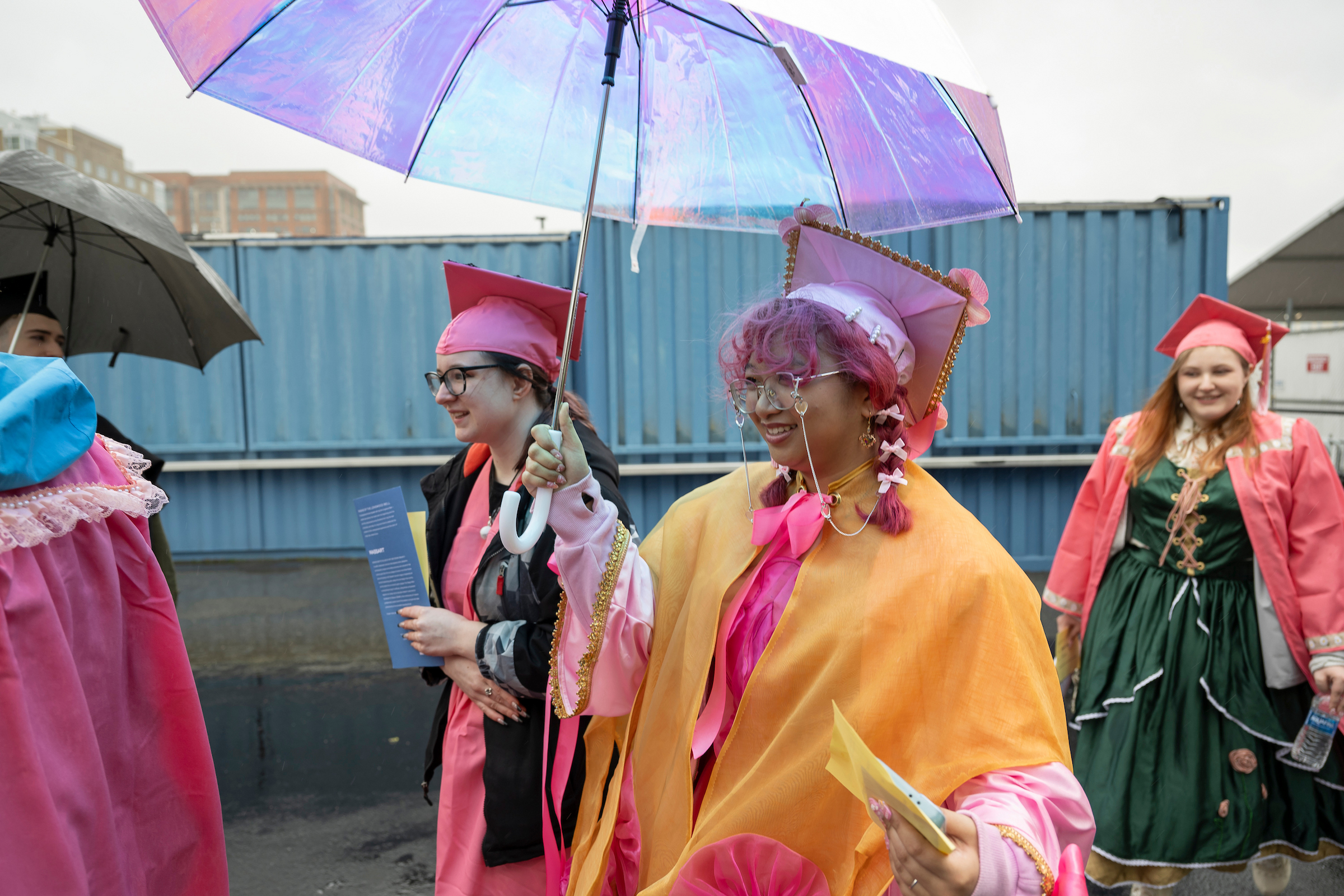 People in colorful, whimsical graduation robes and caps stand outside, some holding umbrellas. One person in the foreground wears a bright pink and yellow outfit with glasses and purple hair, smiling during the commencement. Shipping containers are in the background.