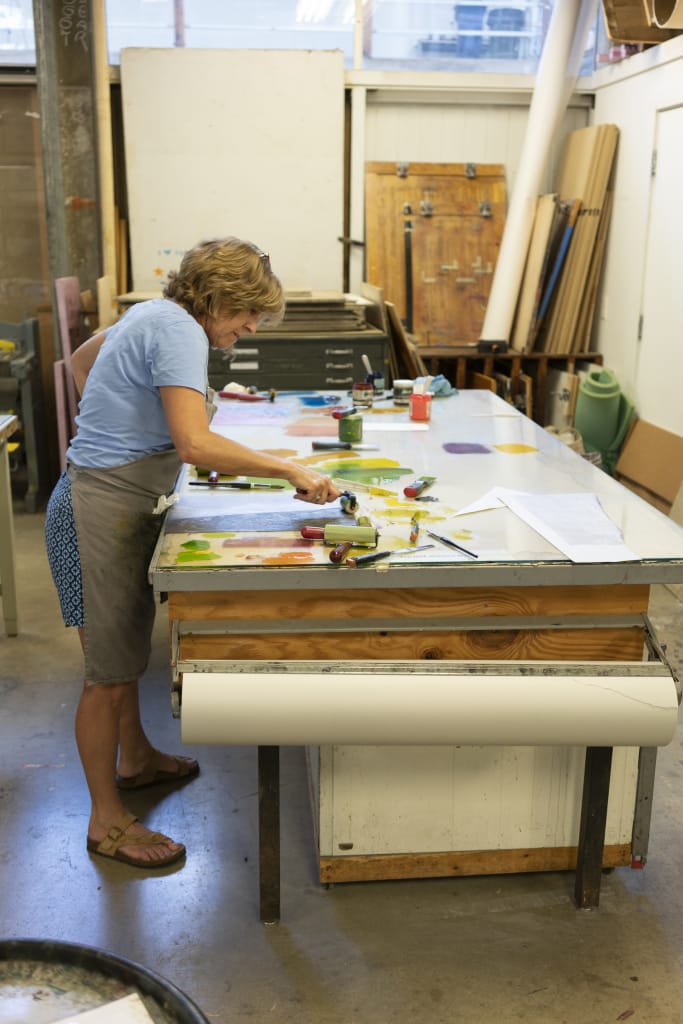 A woman in a blue shirt and gray skirt works at a large, cluttered table in an art school studio, using a roller and colorful paints. Art supplies, paper, and tools are scattered around the creative New England workspace.
