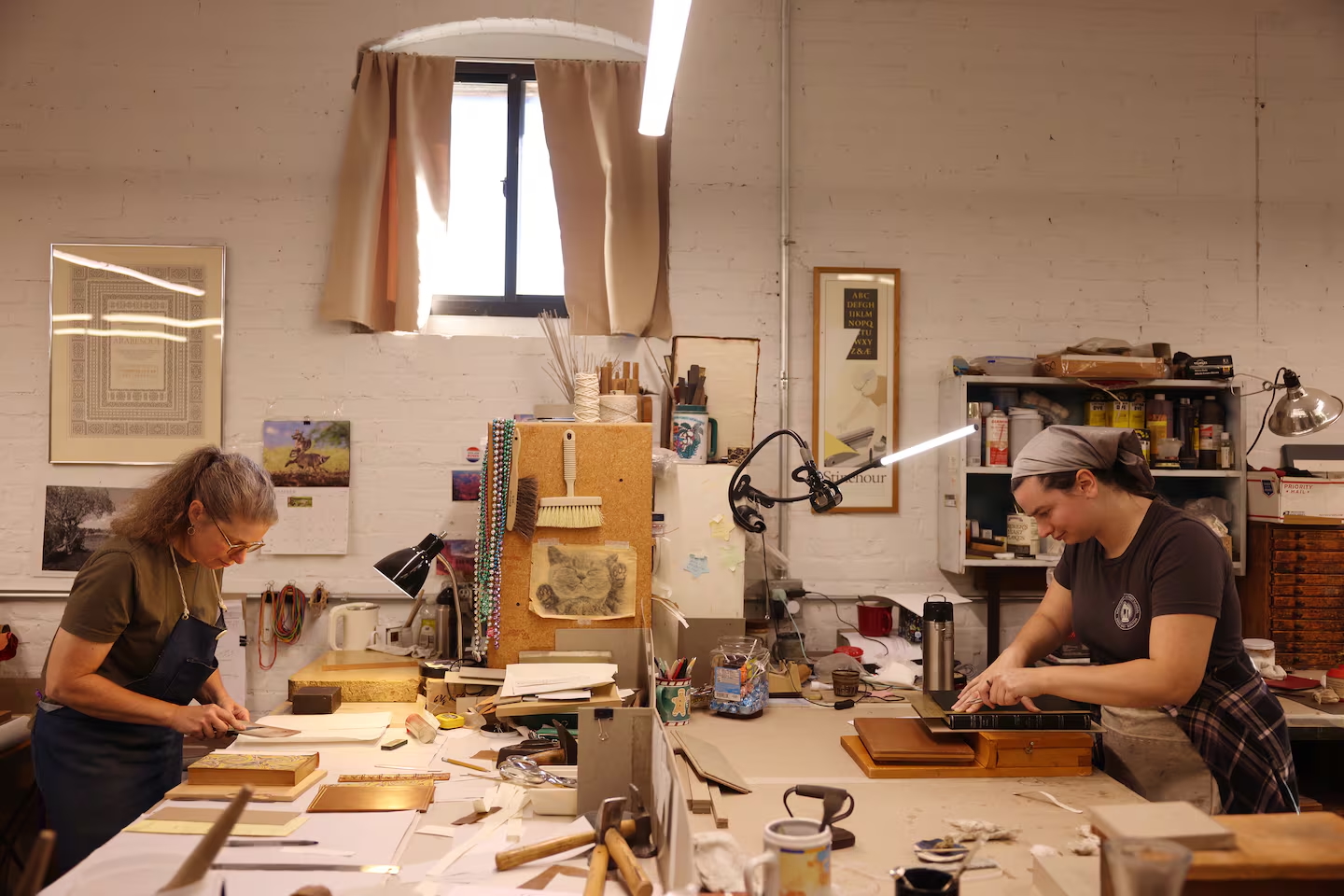 Two women, including MassArt Alum Francesca Santiago, work at separate tables in a well-lit, organized workshop surrounded by tools and craft materials. Shelves and art supplies line the walls, creating a creative and focused atmosphere reminiscent of Harcourt Bindery.
