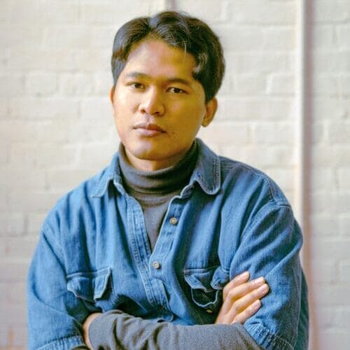 A person with short, dark hair sits with arms crossed, wearing a denim shirt over a gray turtleneck. Set against a light brick wall, the individual—possibly Sopheak Sam of MassArt and Boston Artadia Award fame—has a calm, neutral expression.