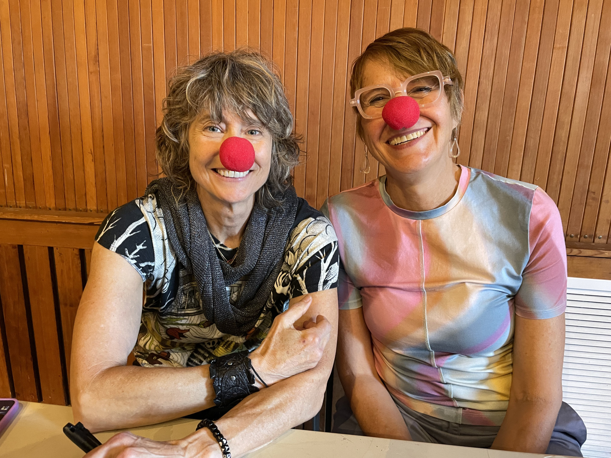 Two smiling women sit side by side at a table, both wearing red clown noses. The background features vertical wooden paneling. One is a MassArt alumna with curly hair, while the other wears glasses and a colorful shirt.