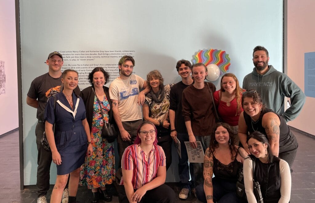 A group of thirteen people pose and smile in front of a blue exhibition wall reading NANCY CALLAN KATHERINE GRAY and CLOWN IN ME LOVES YOU at Fuller Craft Museum, celebrating work by MassArt Alumna Nancy Callan at the art gallery.