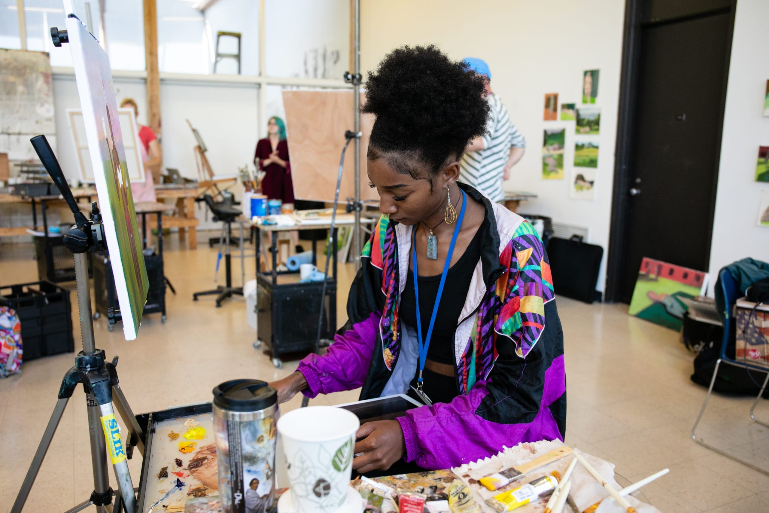 A person wearing a colorful jacket sits at a table in a vibrant MassArt studio, focusing on their work. Paints, brushes, cups, and art supplies are scattered around, with other people and artworks visible in the background.