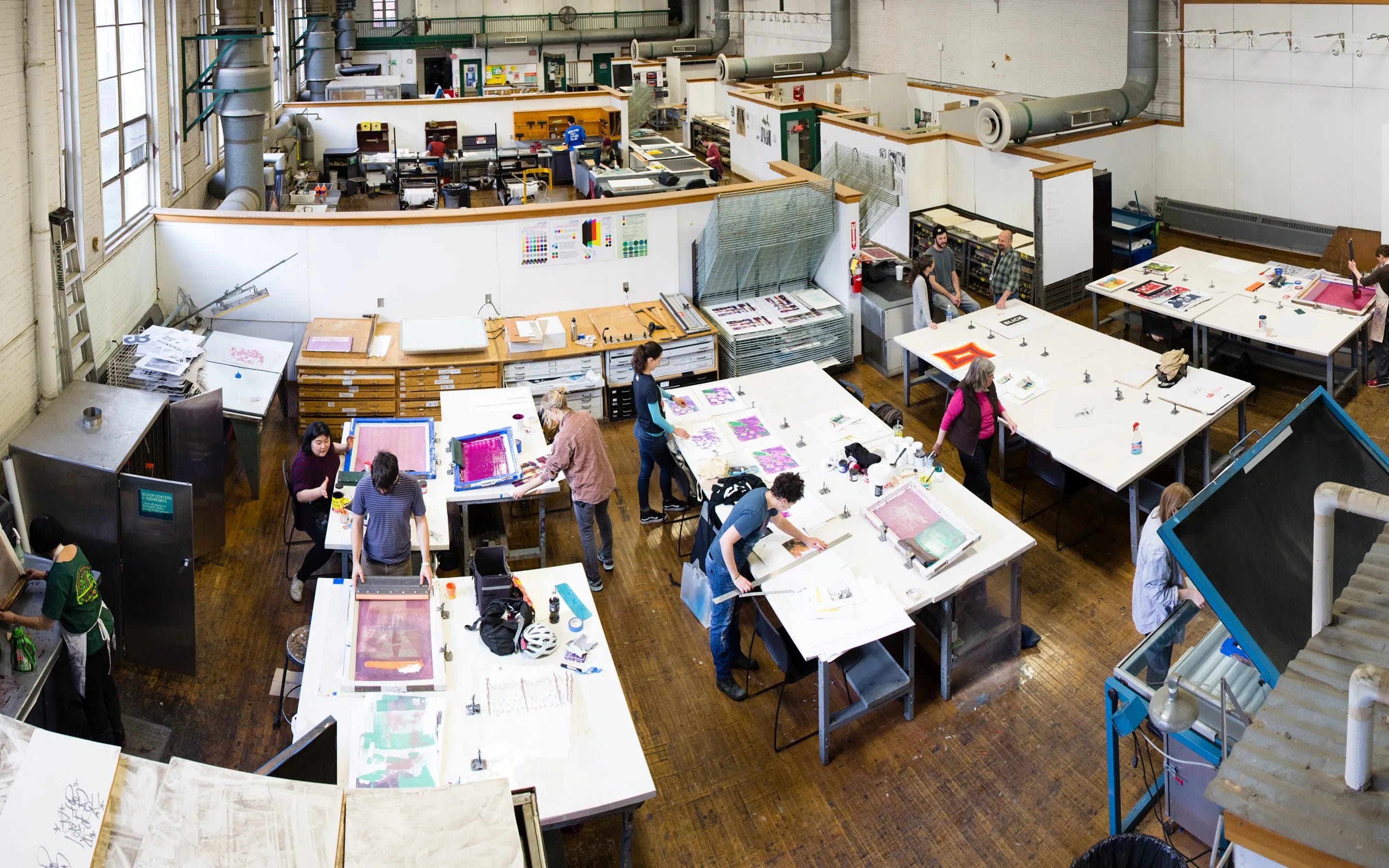 A bright, spacious art studio with several people working at large white tables on various creative projects as part of Continuing Education. Art supplies and colorful materials are scattered around, with printing equipment visible in the background.