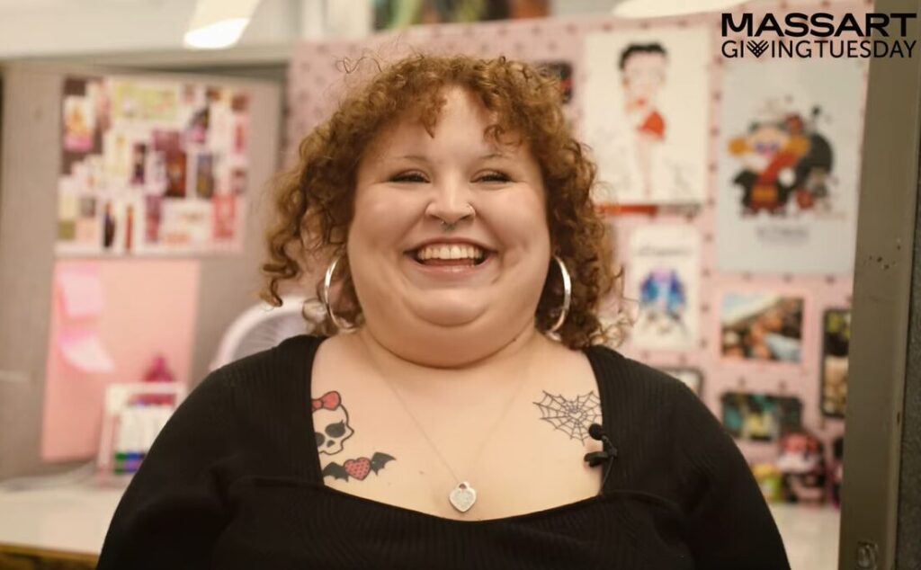 Natalie Morris, a MassArt student, smiles in an art-filled workspace. With curly hair, hoop earrings, and tattoos of a spiderweb and skull with a bow, her student story shines. MassArt Giving Tuesday is in the corner.