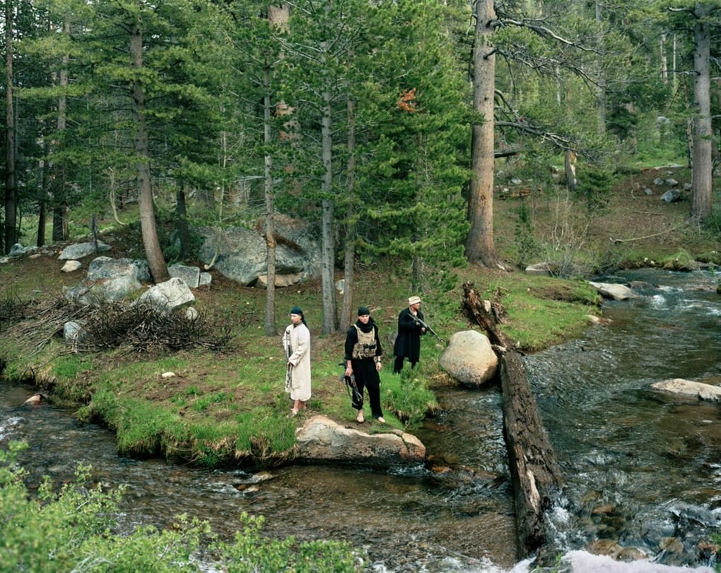 Three people stand on rocks beside a flowing creek in a forest, surrounded by tall trees and greenery. MassArt alum Claire Beckett, known for her work exploring military training, captures the moment as water rushes over nearby rocks.