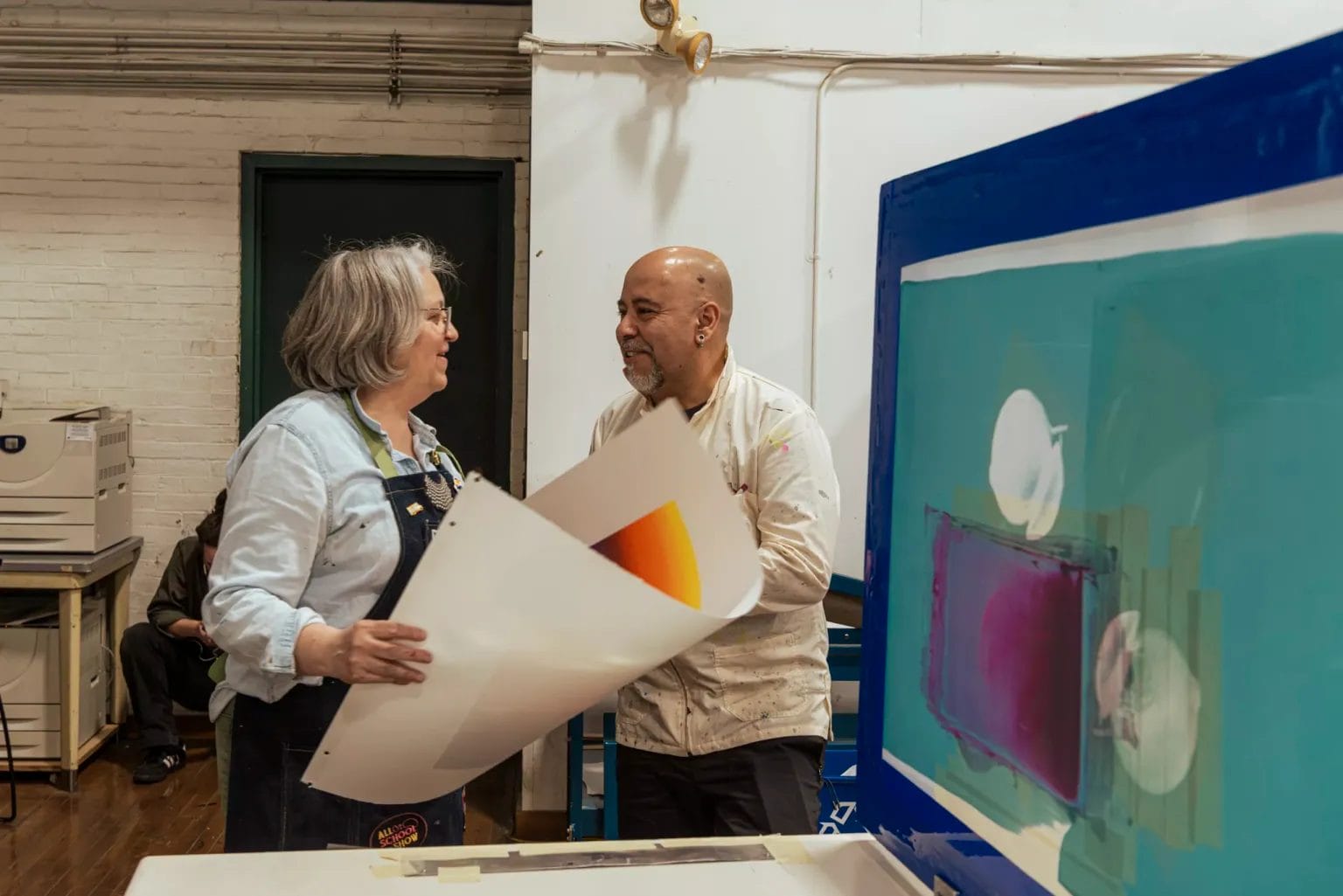 Two people stand in an art studio, holding and discussing large colorful prints. One person wears an apron, and abstract artwork is visible on a table and an easel beside them.