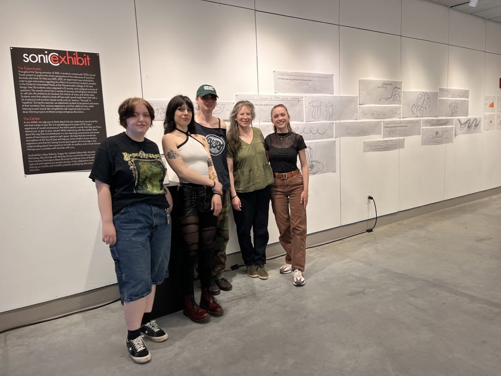 Five MassArt students and alumni stand side by side in front of a gallery wall displaying pencil sketches and a sign titled “sonicxhibit.” The group appears relaxed, posing for a photo in the well-lit Science of Sound exhibition space.