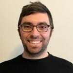 A man with short brown hair, glasses, and a beard is smiling at the camera. He is wearing a black sweater and standing in front of a plain light-colored background, embodying the approachable style of innovation advisors.