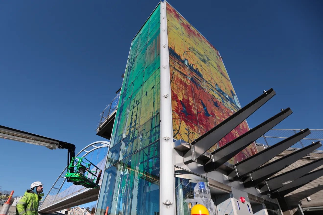 A worker in safety gear stands near a tall glass art structure by Tracy Silva Barbosa with colorful abstract designs, next to a modern pedestrian bridge under a clear blue sky.