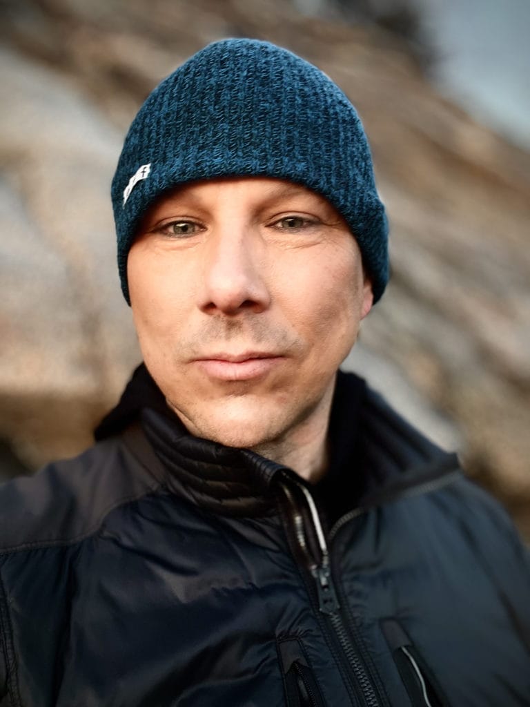 A person wearing a blue knit beanie and a black jacket is standing outdoors in front of a large, blurred rock formation. The lighting is soft, and the person is looking directly at the camera with a slight smile.