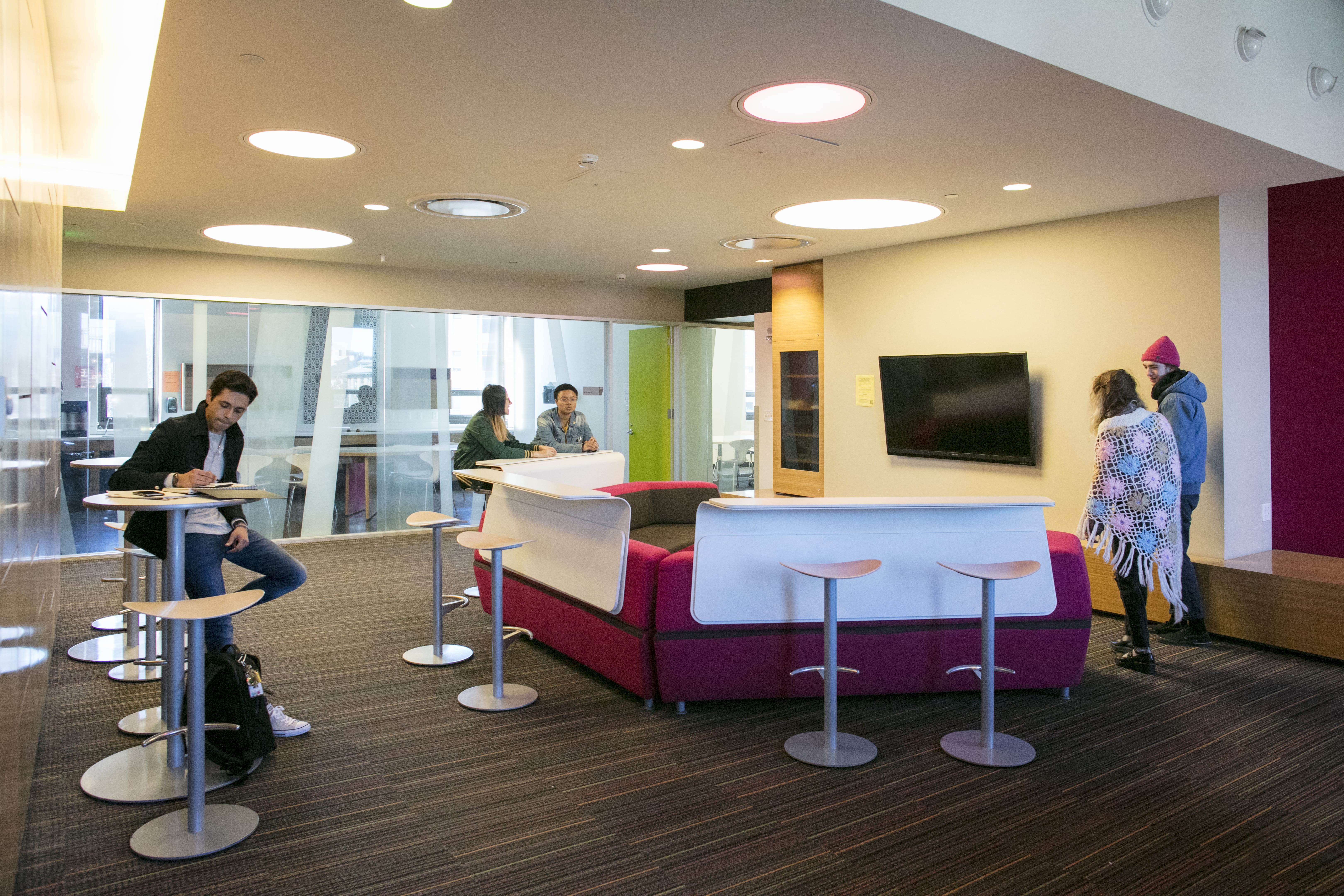 A modern lounge with people studying and talking. There are tall tables with stools, a red couch facing a TV, and large windows letting in natural light—a casual spot to chat about housing costs or meal plan costs in a relaxed, social atmosphere.