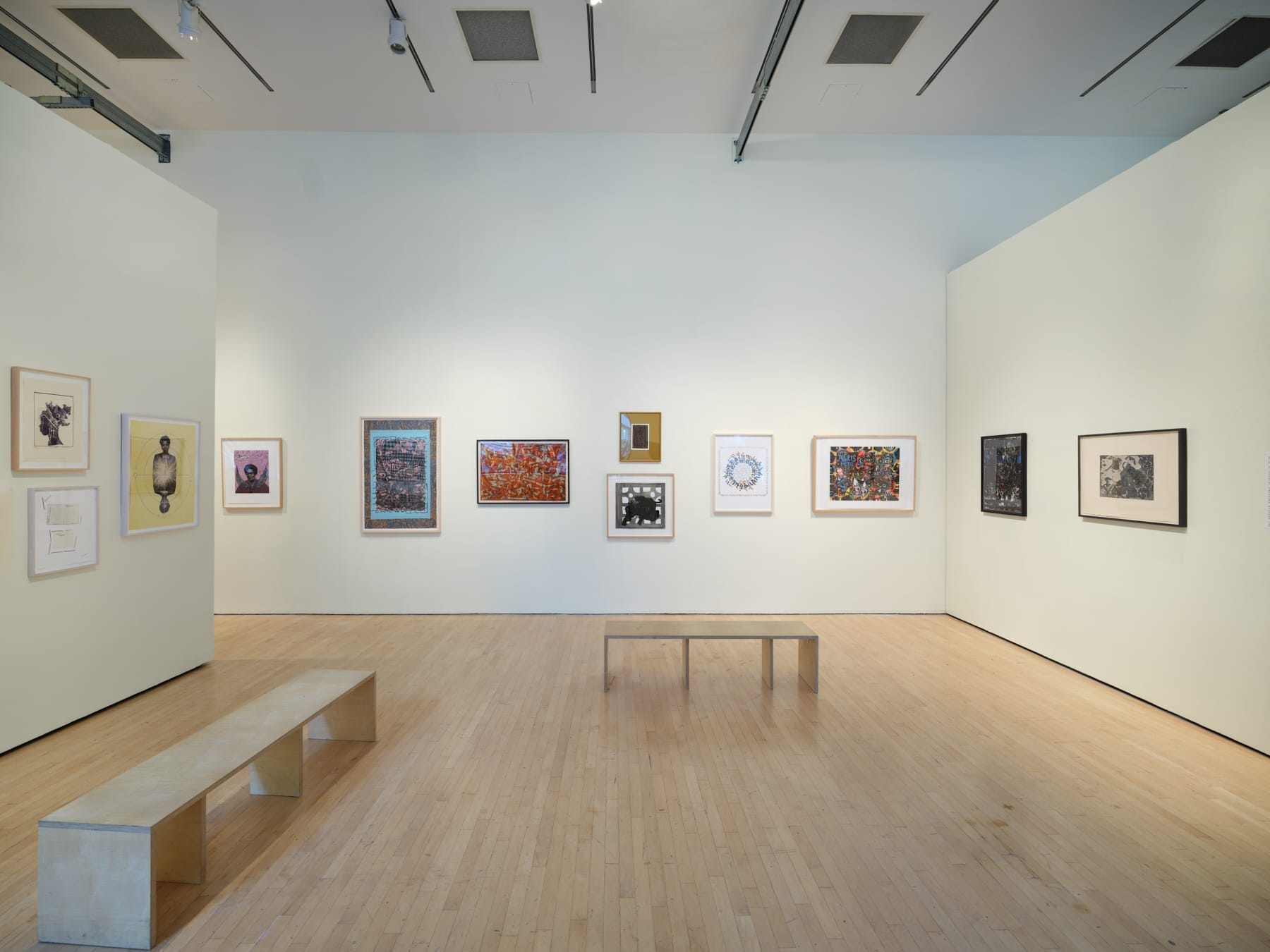 A modern art gallery featured showcases colorful artworks and photographs on white walls. Two simple wooden benches invite visitors to sit and discover new things to do in this spacious, well-lit room with light wood floors.