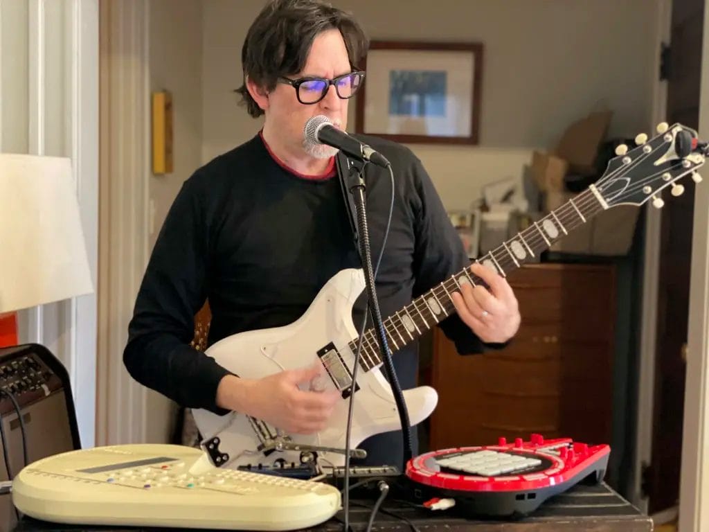 A person with dark hair and glasses plays a white electric guitar and sings into a microphone. Electronic music equipment is set up in front of them on a table in a cozy room.