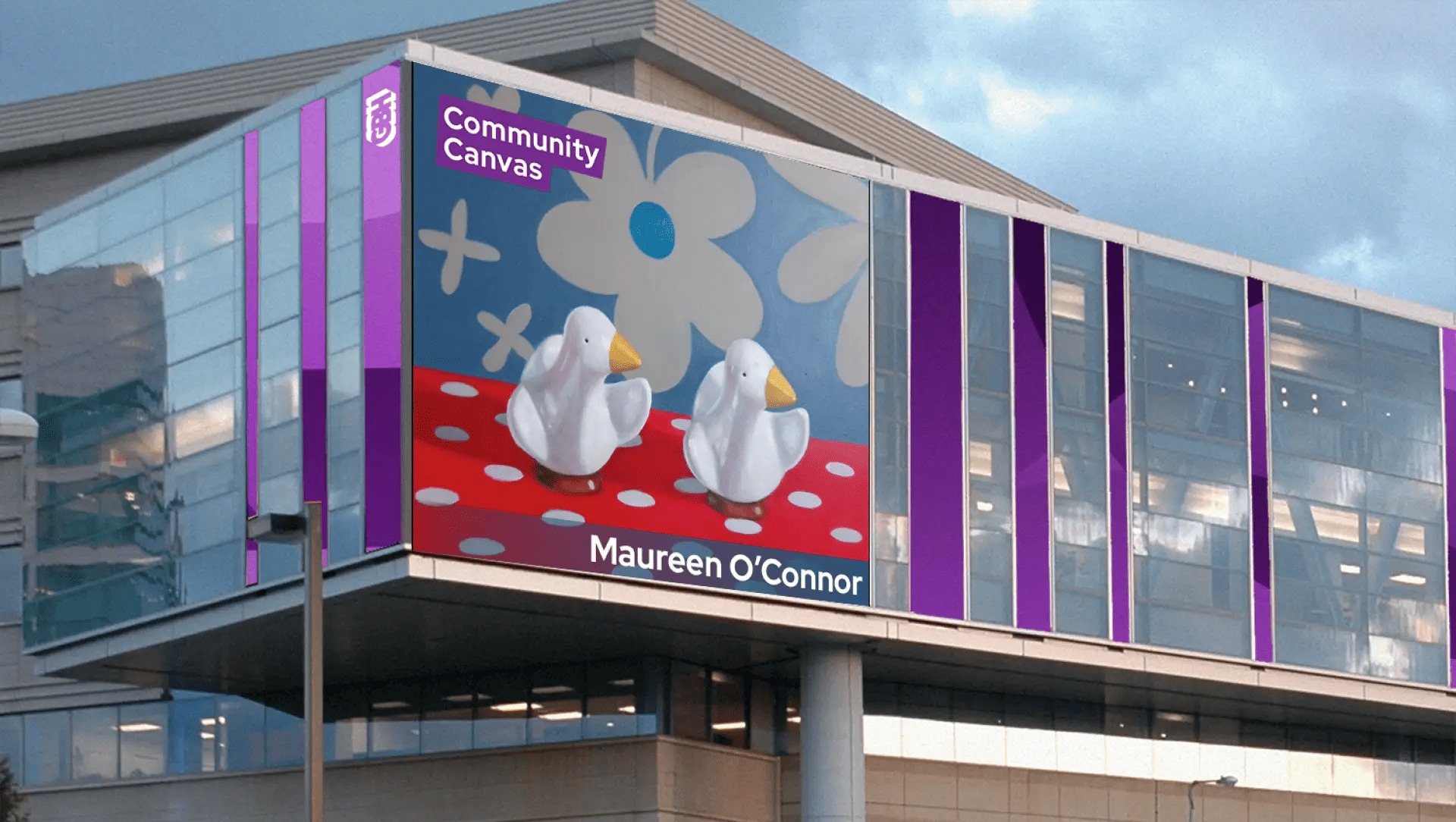 A large digital billboard on a modern building displays two white duck figures with yellow beaks on a red polka-dot surface, with a blue background featuring white flowers.