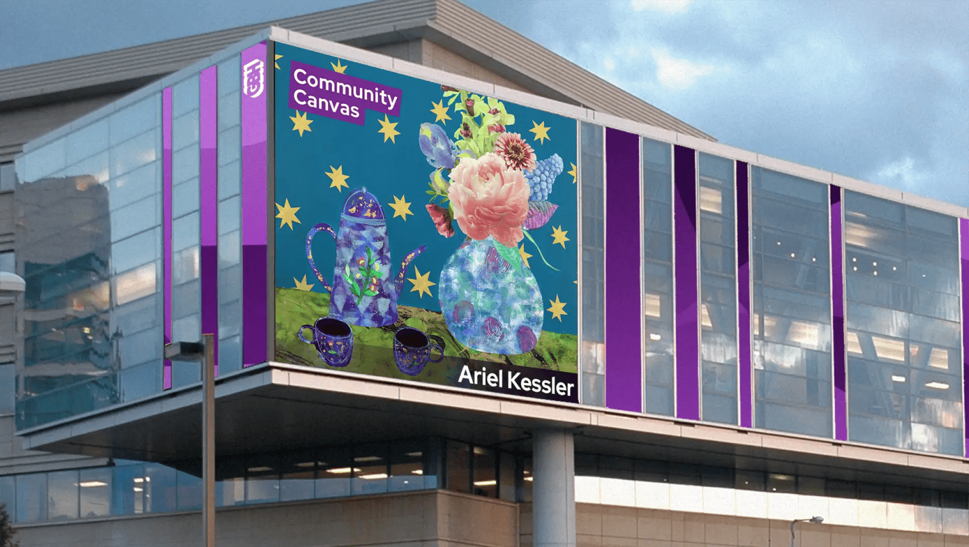 A large billboard on a modern building displays colorful artwork by MassArt Alum Ariel Kessler, featuring a teapot, cups, and floral bouquet on blue with yellow stars. The text reads GBH Community Canvas and Ariel Kessler.