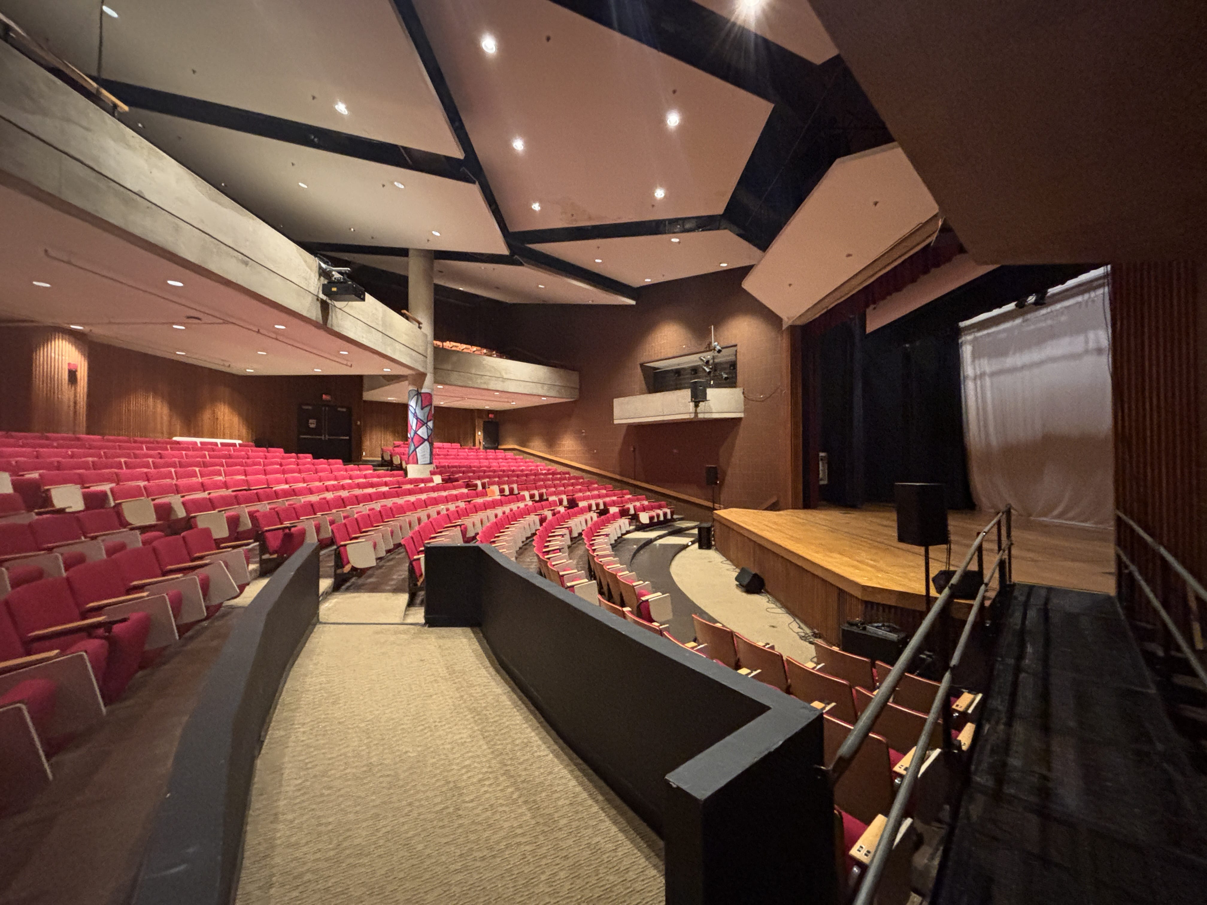 A spacious auditorium with rows of red seats facing a wooden stage, curved seating arrangement, modern ceiling lights, and a carpeted aisle—ideal for hosting Event Services or Conference Services.