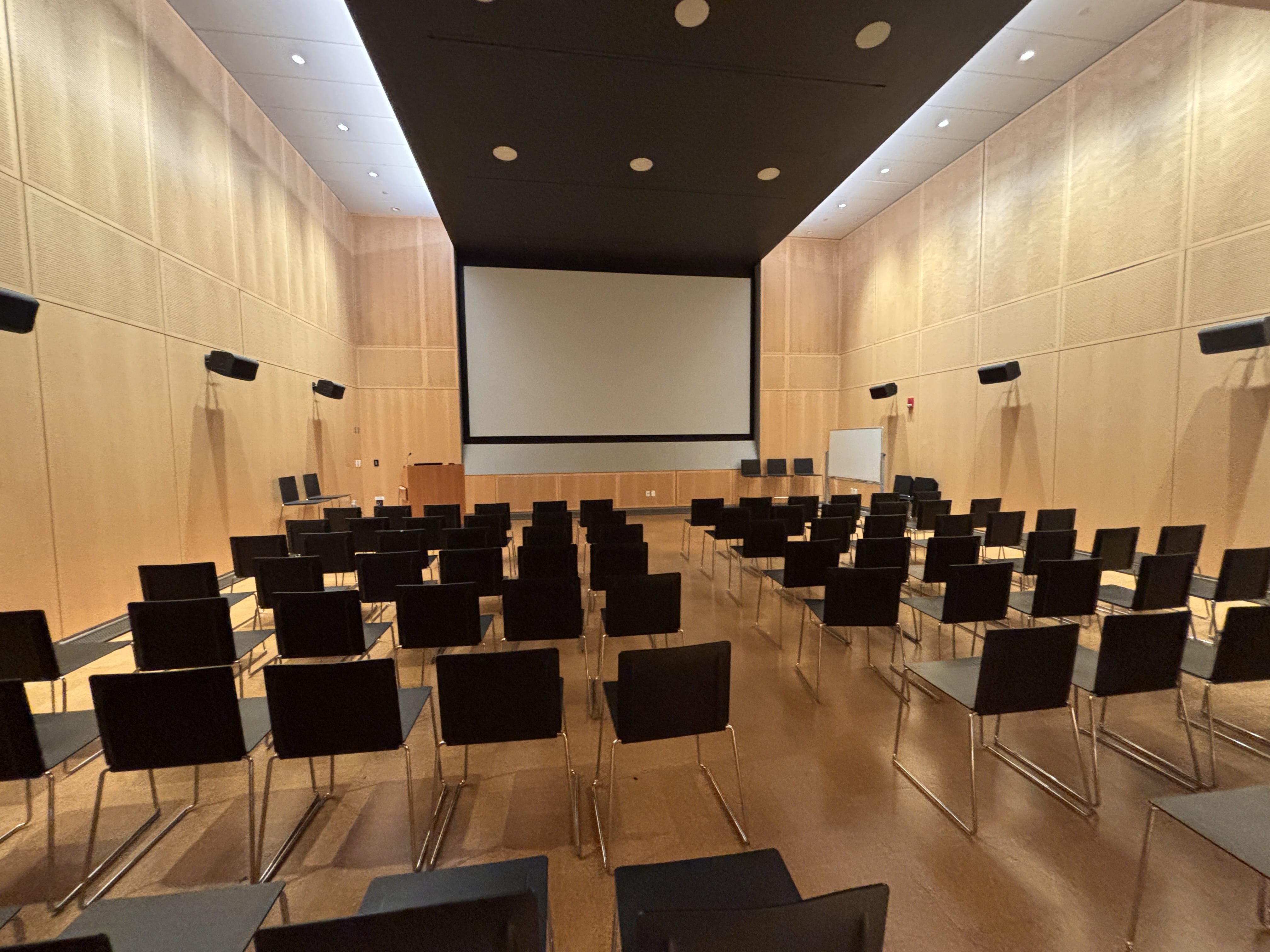Rows of empty black chairs face a large projector screen in a modern, wood-paneled lecture hall with high ceilings and wall-mounted speakers—an ideal setting for professional event services or conference services.