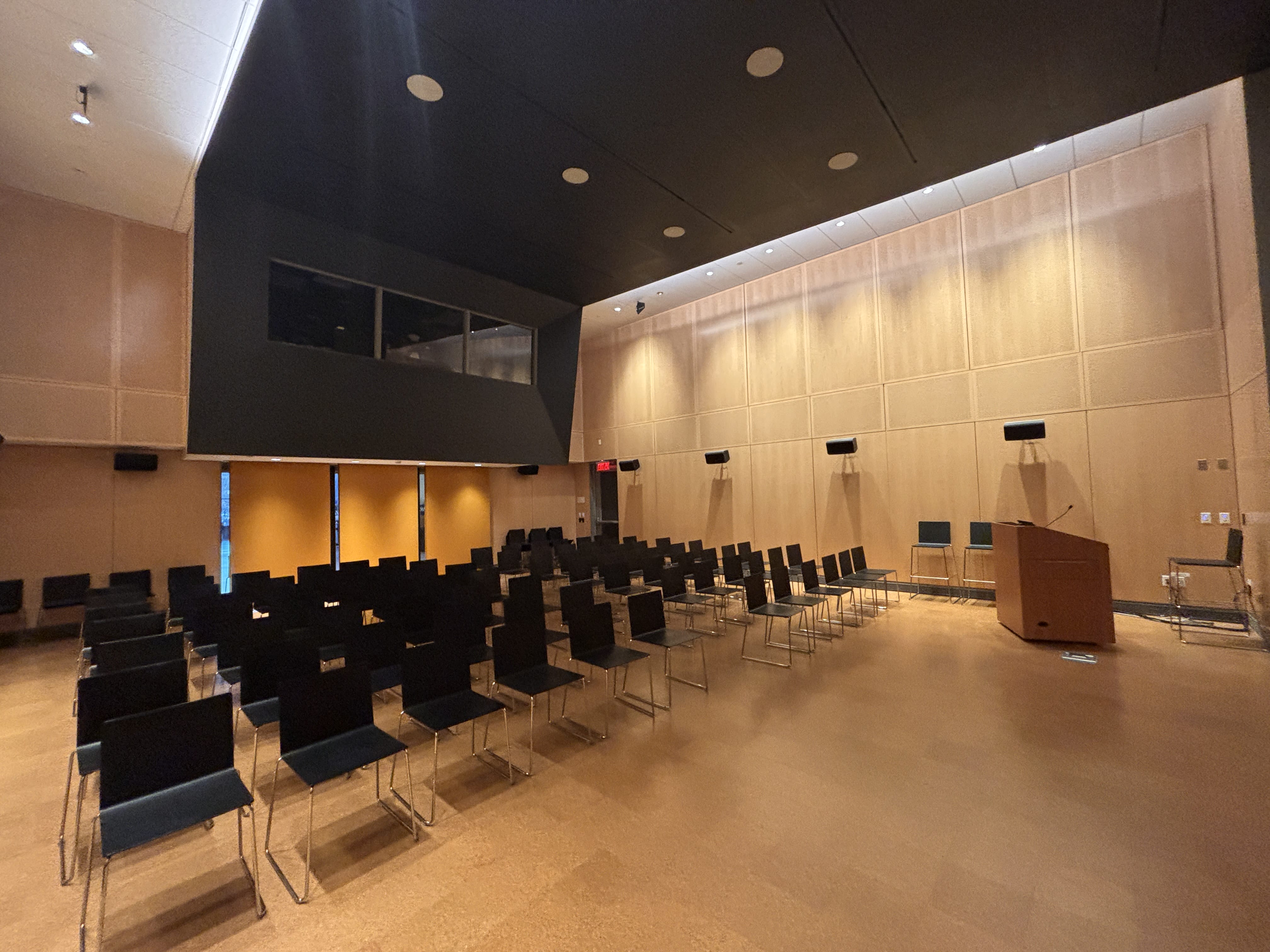A modern lecture room with rows of empty black chairs facing a wooden podium on the right, wood-paneled walls, a cork floor, and a large windowed booth—ideal for conference services. The room is well-lit with ceiling lights.