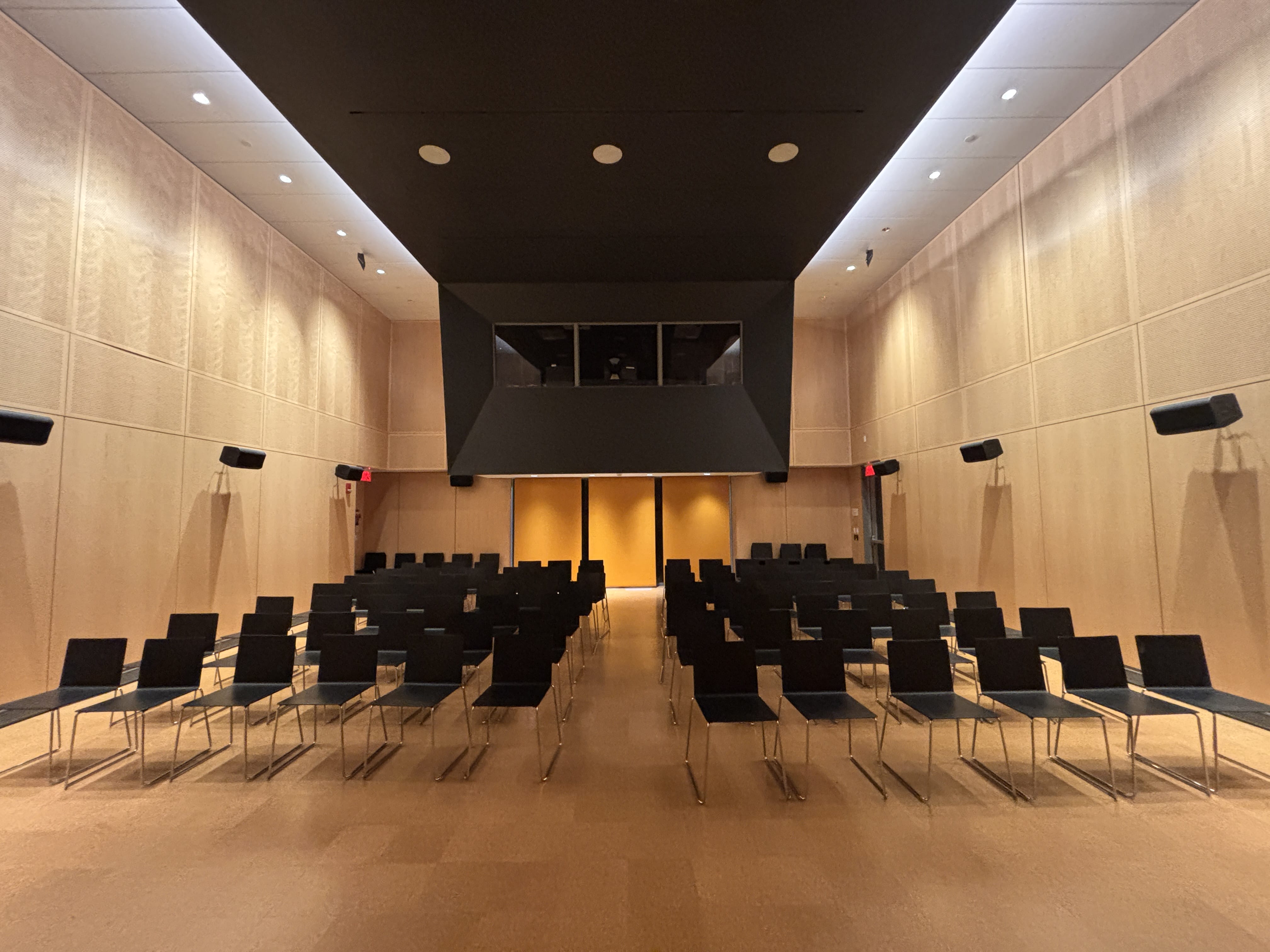 A modern, empty auditorium with rows of black chairs facing a stage and control booth, ideal for conference services. Light wood-paneled walls and a high ceiling with recessed lighting create an inviting event space.