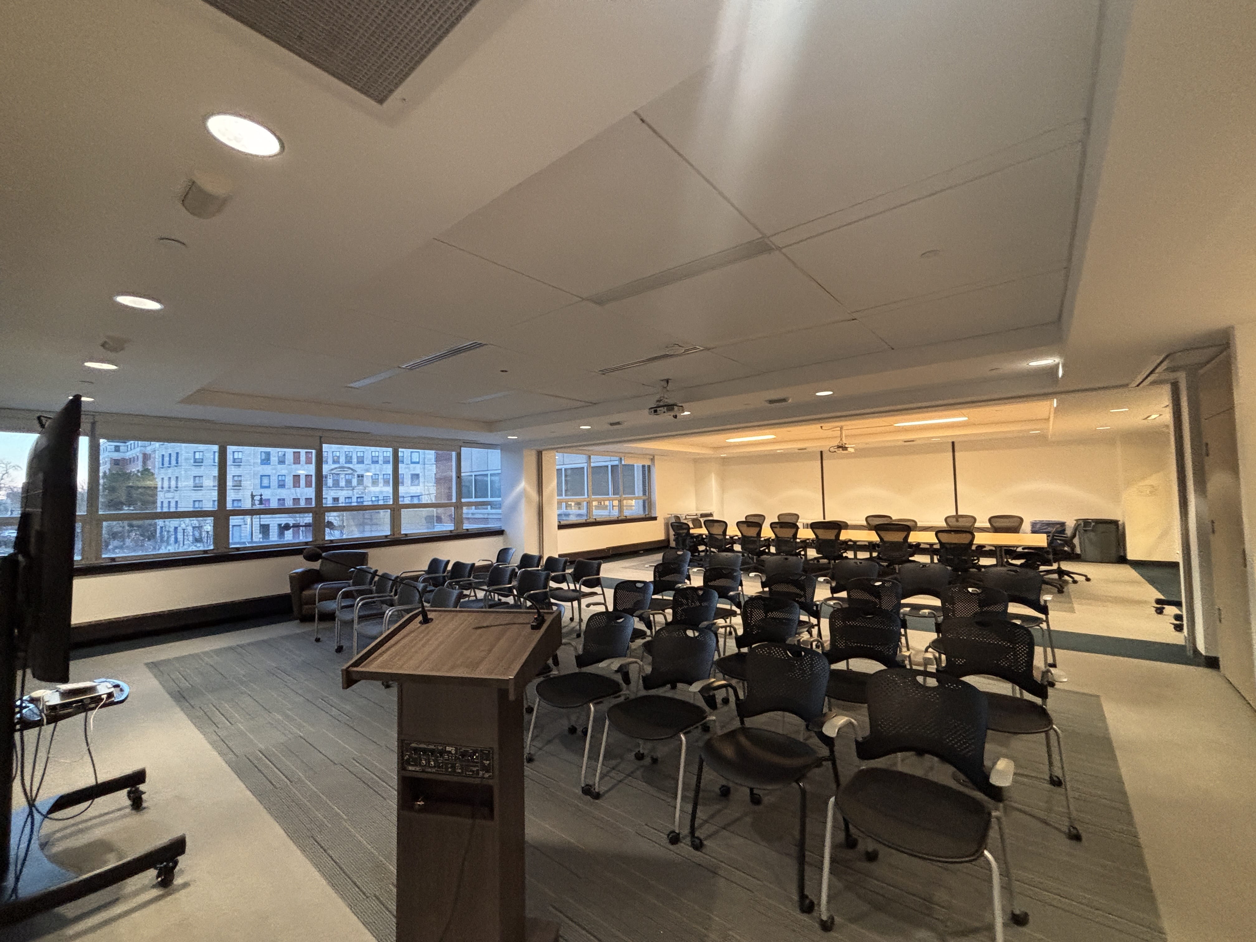 A modern conference room with rows of empty black chairs facing a podium and large TV screen, perfect for event planning or conference services, with windows along one wall letting in natural light and city buildings visible outside.