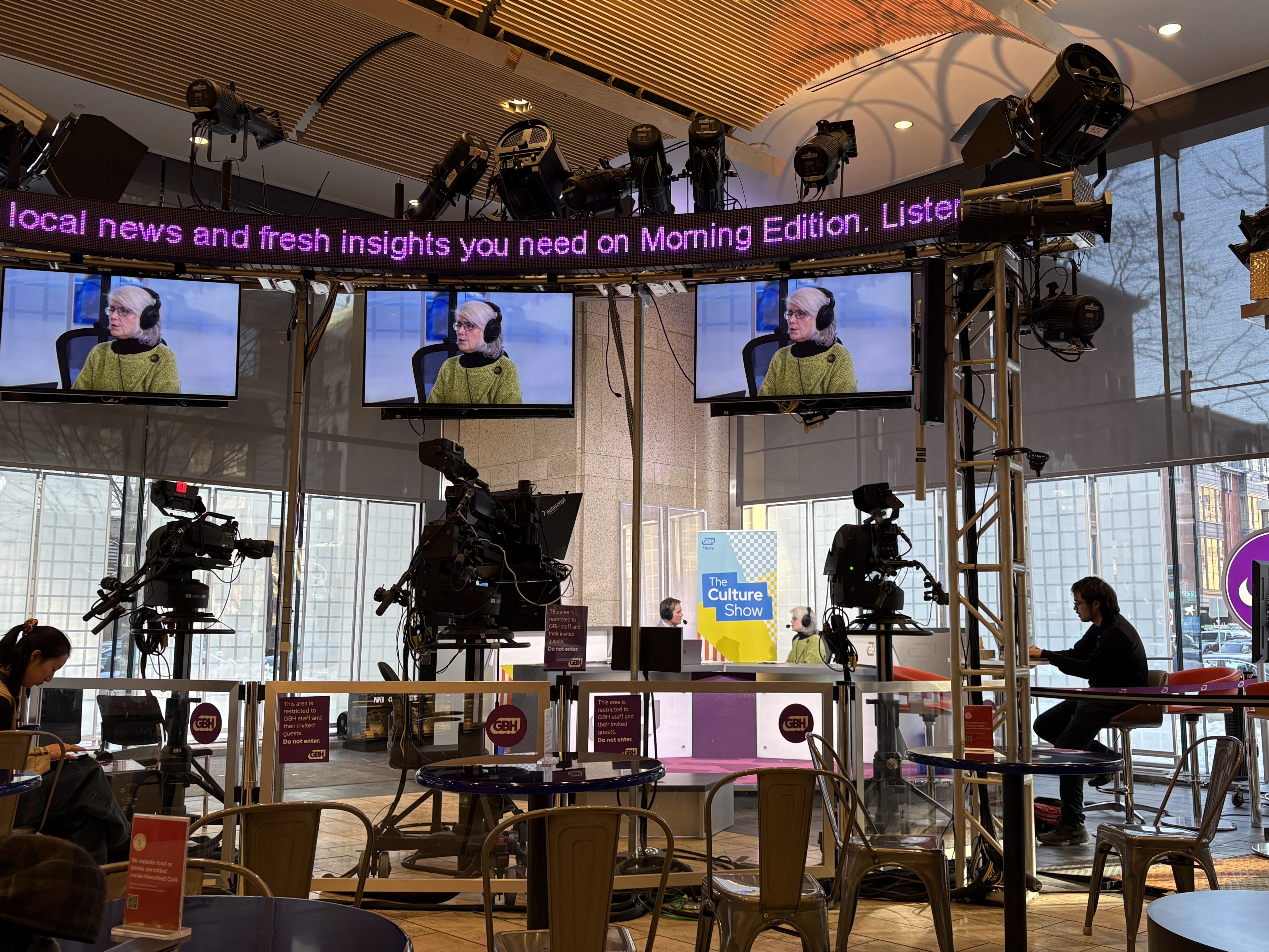 A modern broadcast studio with large cameras, TV screens showing a woman speaking, and a circular news ticker. People at tables discuss Museum Collections on “The Culture Show.” Large windows let in daylight as NPR signs are displayed.