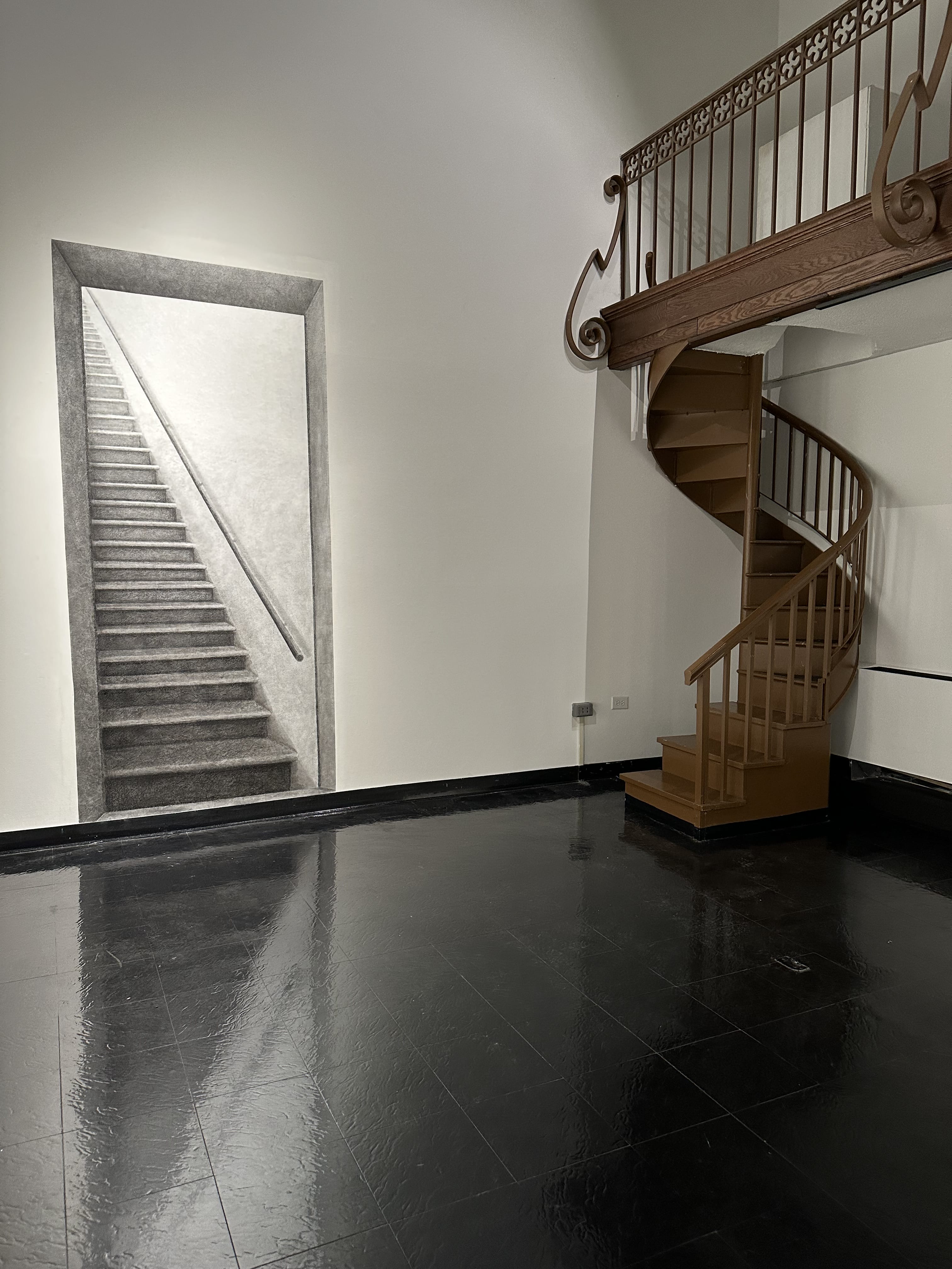 A spiral staircase with a wooden railing stands beside a wall featuring a realistic black and white drawing of a staircase ascending into an open doorway—an artistic touch reminiscent of MassArt New England. The floor is glossy and black.