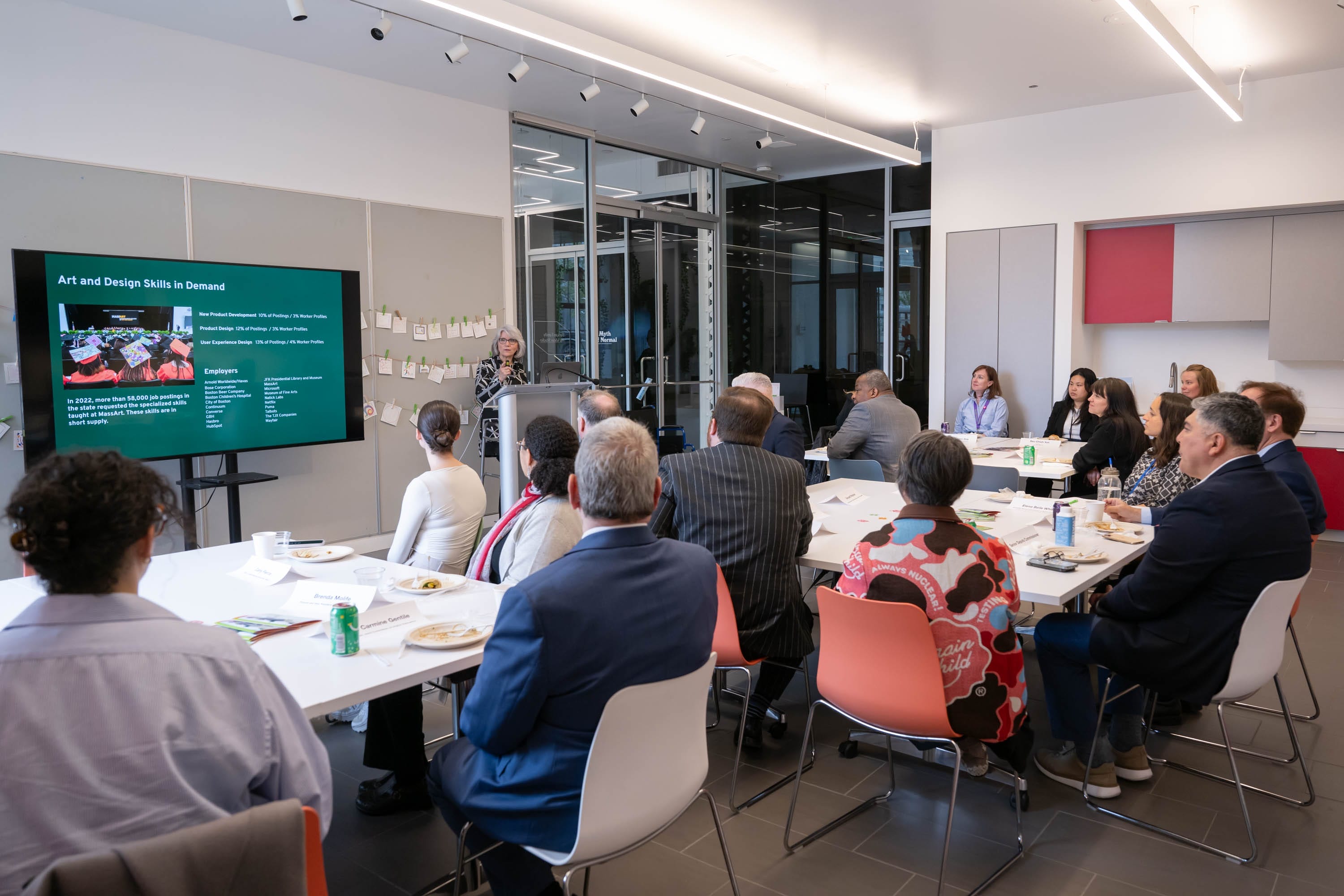 A group of people sit around tables, listening to a speaker presenting a slideshow titled Art and Design Skills in Demand in a modern conference room with glass walls, bright lighting, and professional event services.