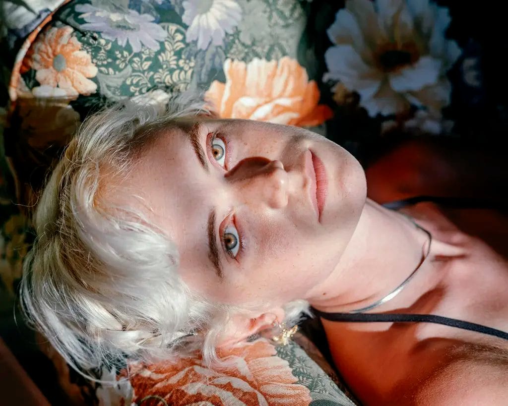 A person with short platinum blonde hair lies on a floral-patterned surface, gazing up into soft, natural light that highlights their face and creates gentle shadows.