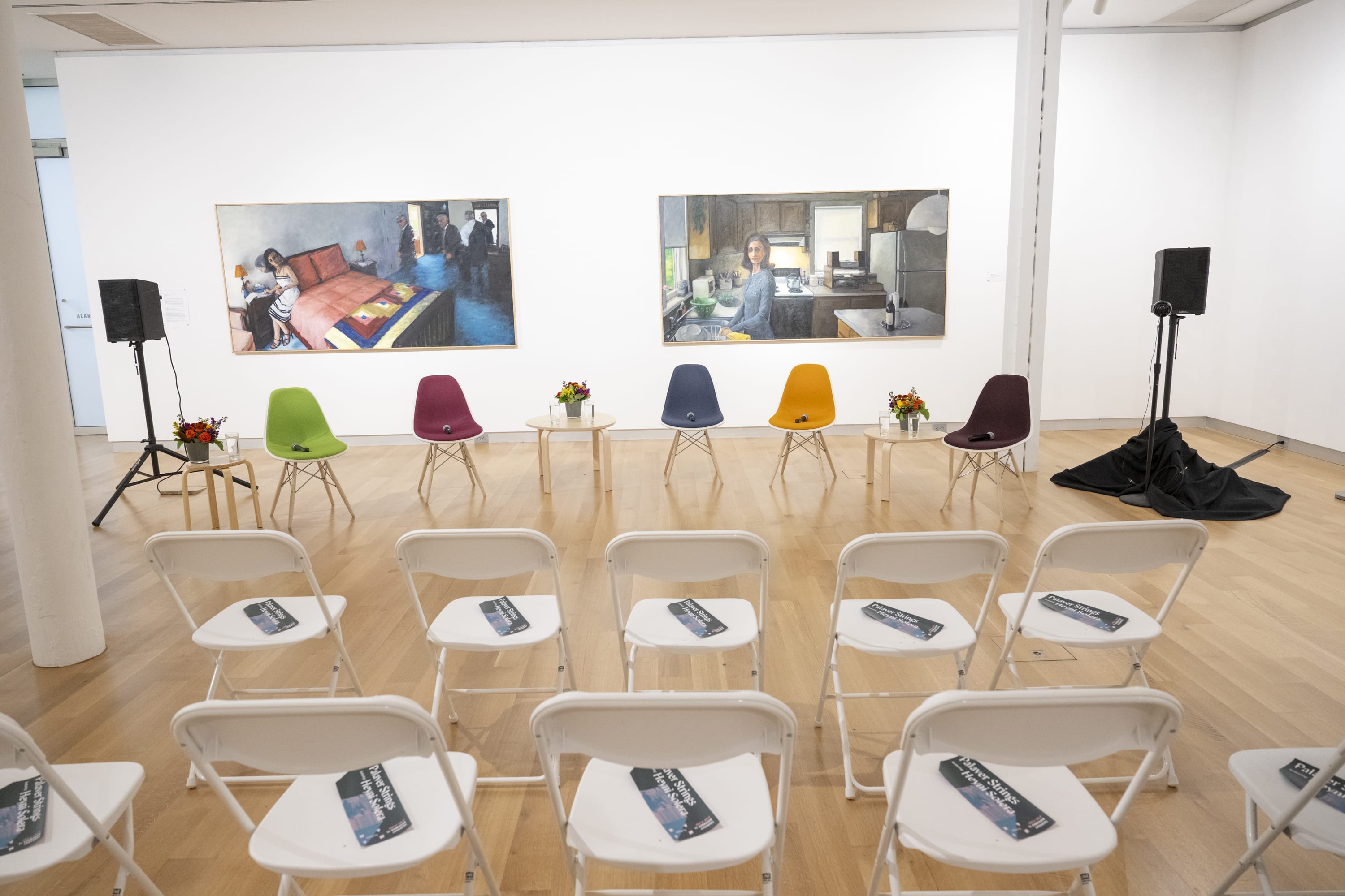 A small gallery setup with six colorful chairs and floral arrangements in front of two large paintings, facing rows of white folding chairs with pamphlets—ideal for conference services or other event services, complete with speakers on either side.