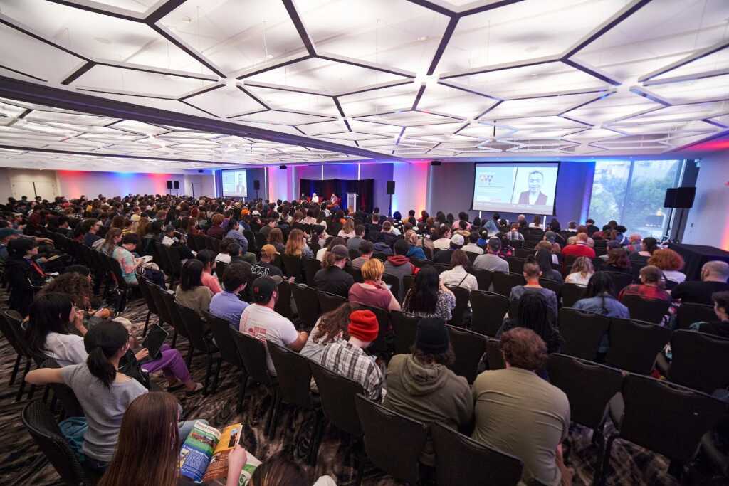 A large audience sits in rows inside a modern conference room, watching a speaker on stage and a presentation projected on large screens at the front of the room.
