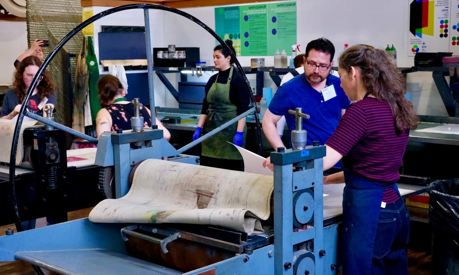 People gather around a large blue printmaking press in an art studio during Alumni Maker Days. Some engage in conversation while others observe or work, surrounded by art supplies, posters, and prints in the background.