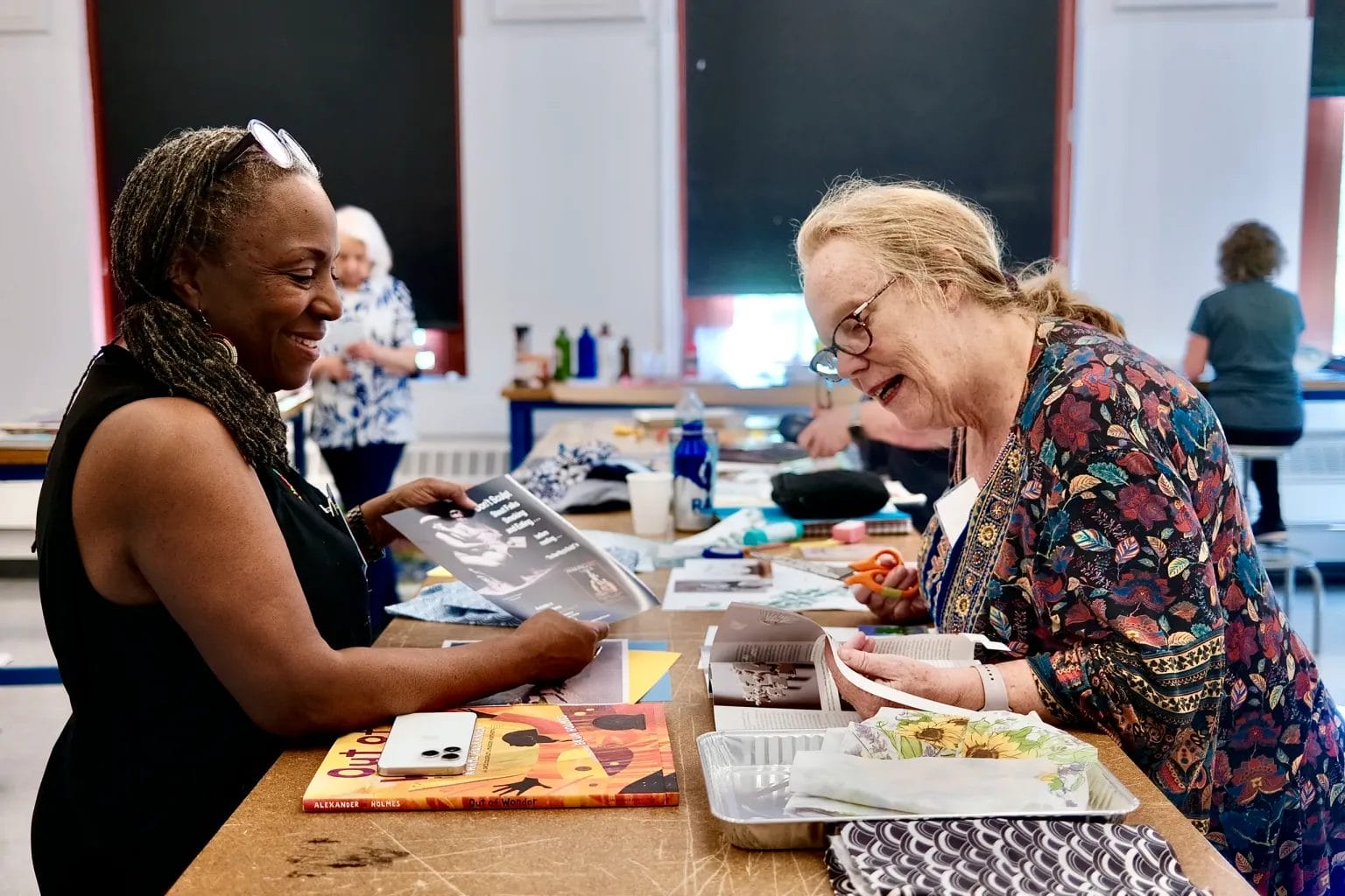 Two women smile and chat while working on an art project at a table during Maker Days, surrounded by books, papers, and supplies. Other alumni and participants are visible in the background of the lively classroom or studio setting.